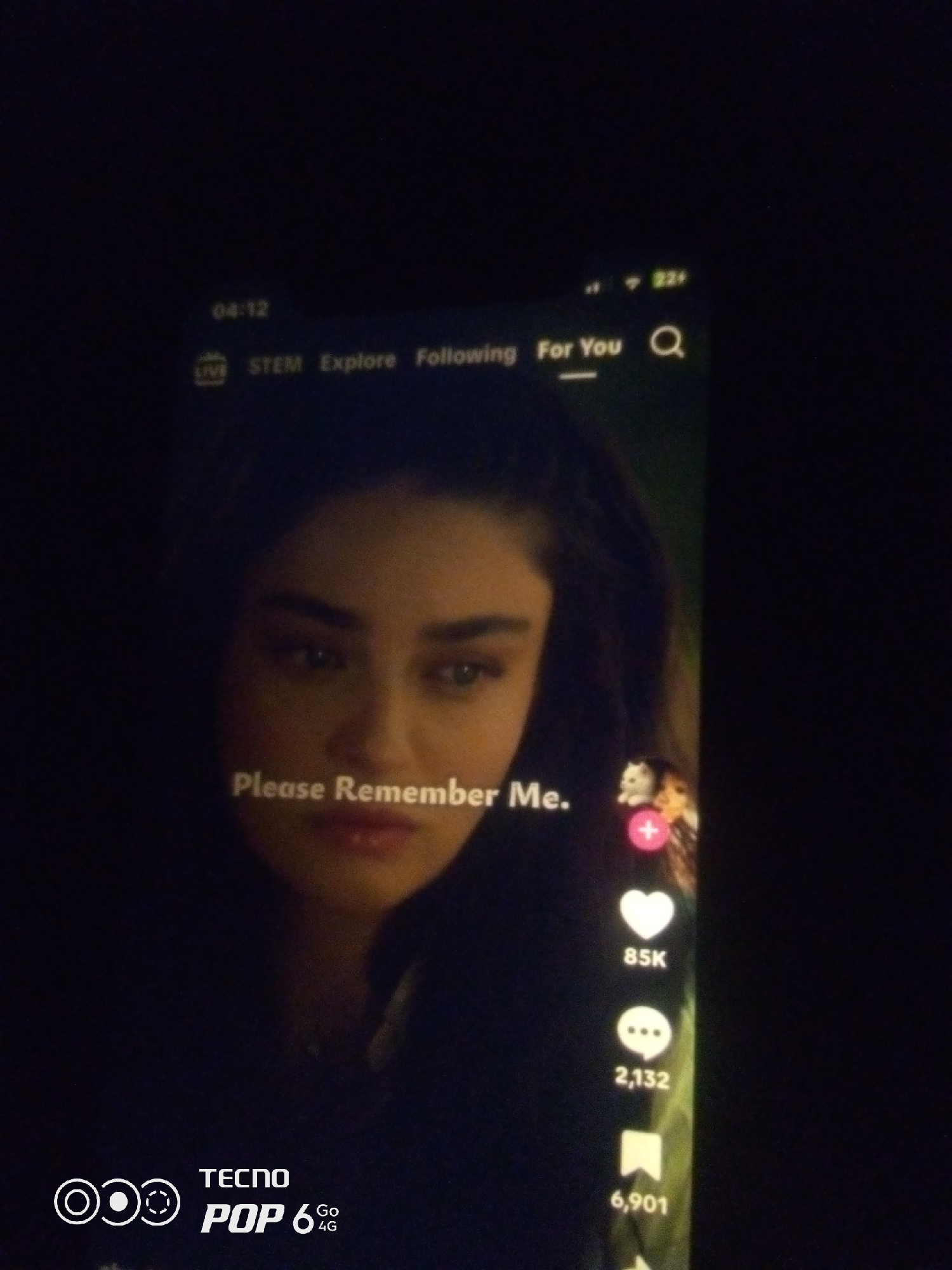 The image shows a close-up of a mobile phone screen displaying a video. The video features a woman with dark hair and brown eyes, her face illuminated by the screen's light. Text overlays on the video read "Please Remember Me.". On the right side of the screen, social media engagement icons are visible: a heart with "85K" below it, a speech bubble with "2,132" below it, and a bookmark icon with "6,901" below it. The top of the screen shows the time "04:12" and various app interface elements. In the bottom left corner, a logo for "TECNO POP 6 Go 4G" is present. The background of the image is dark, suggesting the photo was taken in low light conditions. The exact location in Howayi, Nigeria, cannot be determined from the image.
