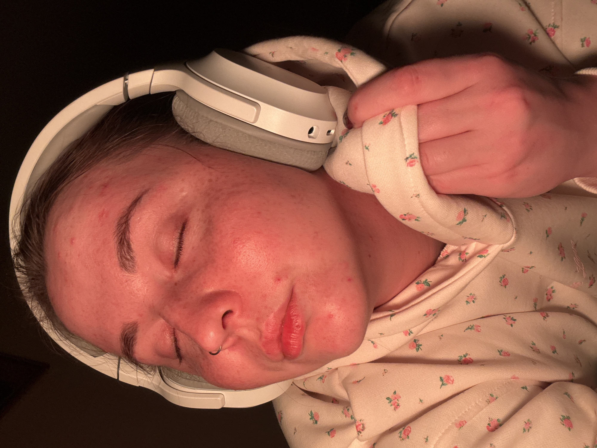 A person with closed eyes and a nose ring is lying down wearing white headphones and a light pink hoodie with a small floral pattern. Their left hand is resting on their chest, holding the sleeve of the hoodie. The background is dark. There is no indication of this image being related to the city of Izhevsk, Russia.
