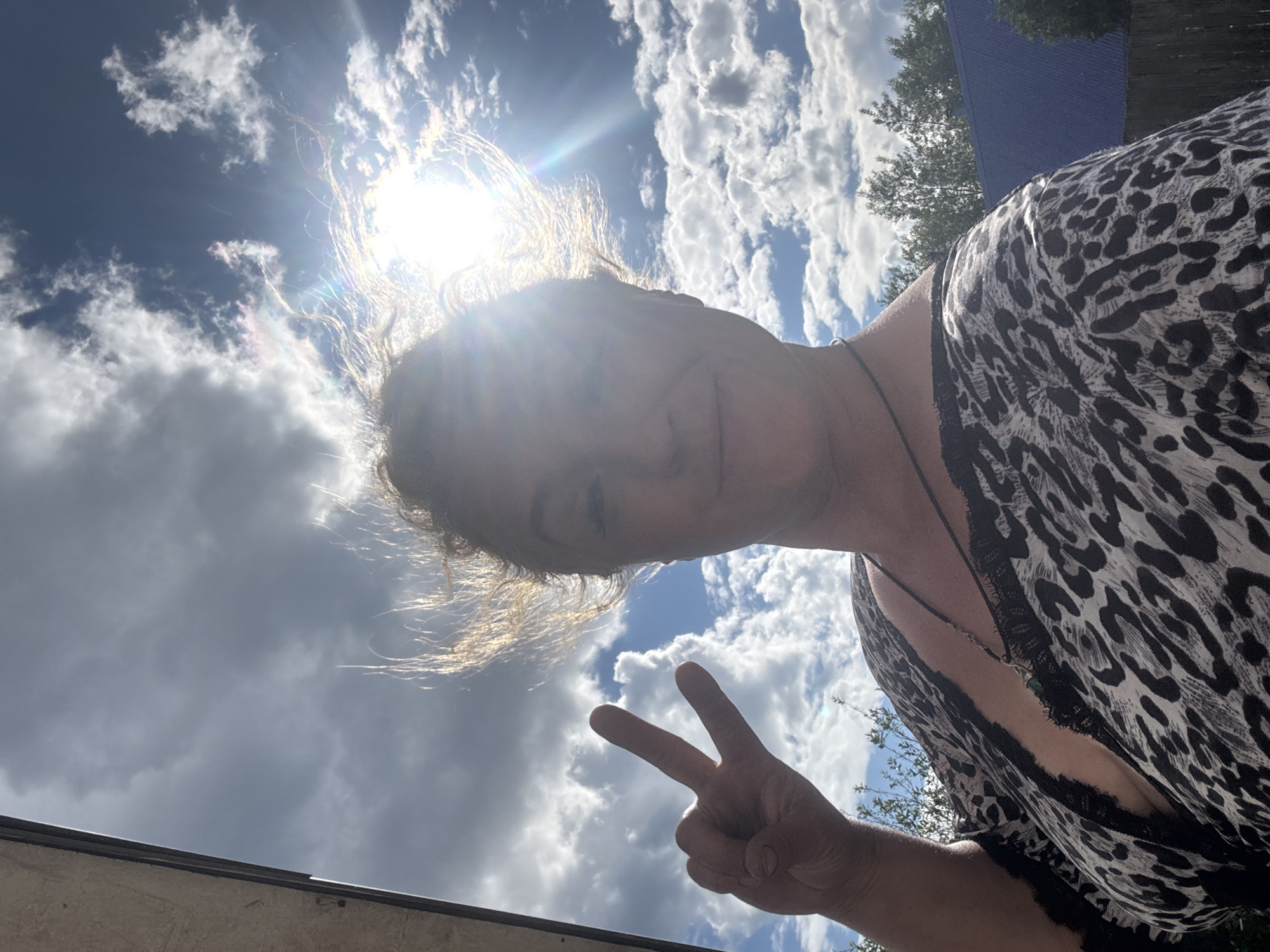 An outdoor, waist-up, selfie-style portrait features a woman with fair skin and blonde hair, backlit by the sun. She is making a peace sign with her left hand, held up to her left side. Her hair is windswept and catches the sunlight. She wears a black and white leopard-print top with black lace trim around the neckline. A dark necklace with a pendant is visible. Behind her, a bright sun shines through scattered clouds, with hints of green trees and a blue roofed structure visible in the background. The scene suggests a casual outdoor setting, possibly in the city of Chaikovsky, Russia, given the weather and vegetation.