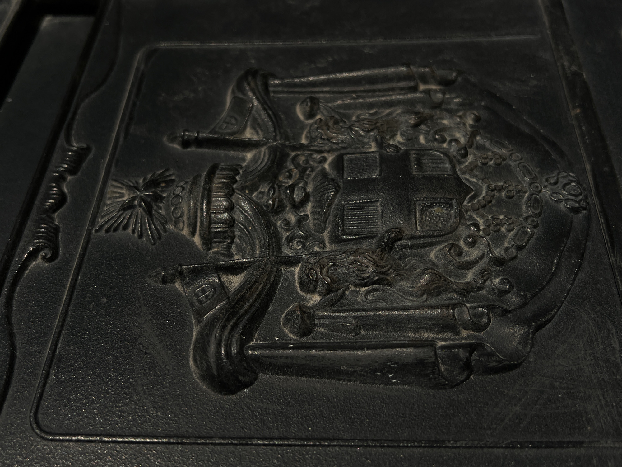 This is a close-up, angled, medium shot of a black, embossed, rectangular plaque. The plaque features a detailed, raised relief carving, likely made of metal or stone, depicting a scene with symbolic elements.
The central subject of the carving is a building with a rectangular structure and a cross on its roof, suggesting a religious building like a church or chapel. To the left of the building is a tall, cylindrical structure topped with a starburst or sun-like emblem, possibly representing a candle or a celestial body. Surrounding the building and other elements are swirling, cloud-like or wave-like forms, with what appear to be dragon heads or mythical creatures integrated into the design. The overall composition evokes a sense of drama and symbolism, possibly representing a significant event or belief.
The lighting is somewhat dramatic, creating shadows that accentuate the raised relief and give the carving a three-dimensional quality. The surface of the plaque appears textured and somewhat worn, hinting at its age or the material used. The edges of the plaque are visible, with a raised border.
There is no visible text within the image. The scene is static, with no apparent activity or event taking place beyond the depiction itself. The location context of Ternopil, Ukraine, does not provide specific visual cues within the image itself, but suggests a potential cultural or historical significance of the artifact.