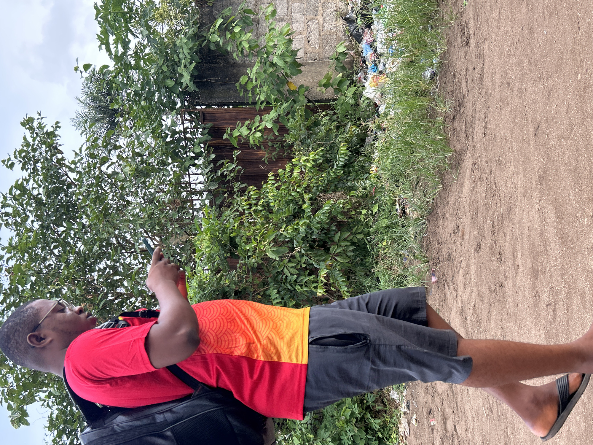 In Ihiagwa, Nigeria, a young Black man wearing a red and orange t-shirt, dark shorts, glasses, and flip-flops walks on a dirt path. He carries a black backpack and is looking down at a mobile phone he holds in his hands. The background features a rusty metal gate, a stone wall, lush green foliage, and a pile of trash. The sky above is overcast.