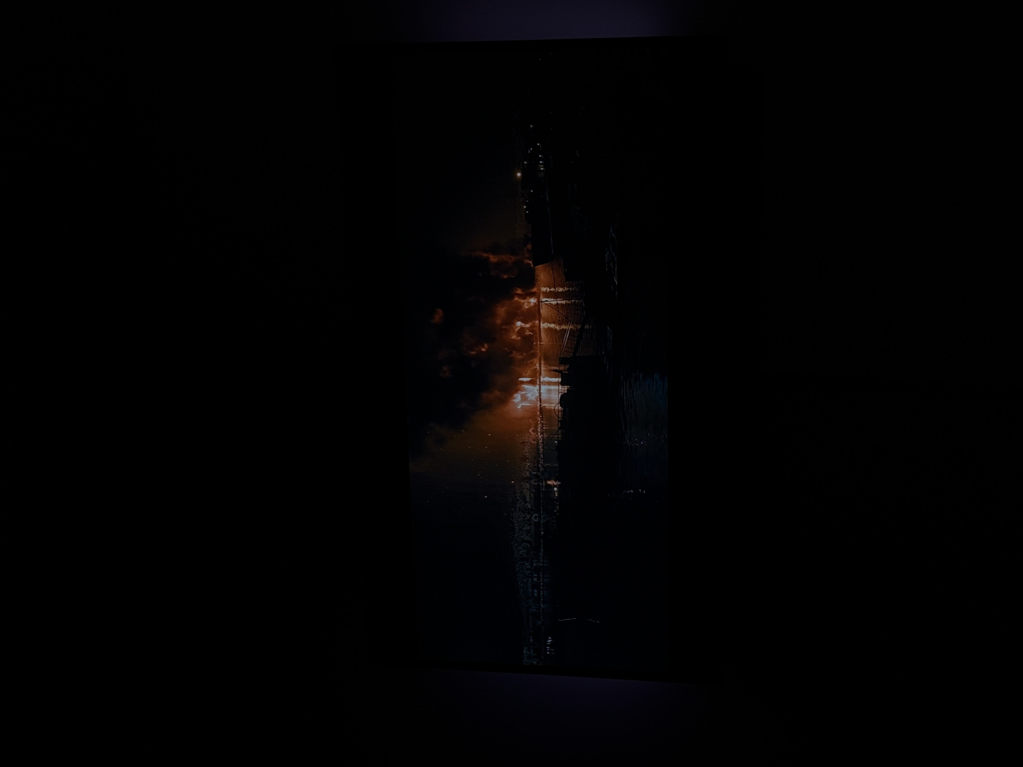 A nighttime scene depicts a significant fire raging on a body of water, likely a river or harbor. Intense orange flames and thick, dark smoke billow upwards, illuminating the water's surface with fiery reflections. Silhouetted structures, possibly buildings or boats, are visible along the shoreline on the left side of the image, with faint lights suggesting an inhabited area. In the foreground, the dark water reflects the fire and some distant lights. The overall impression is one of destruction and chaos in an urban setting at night.