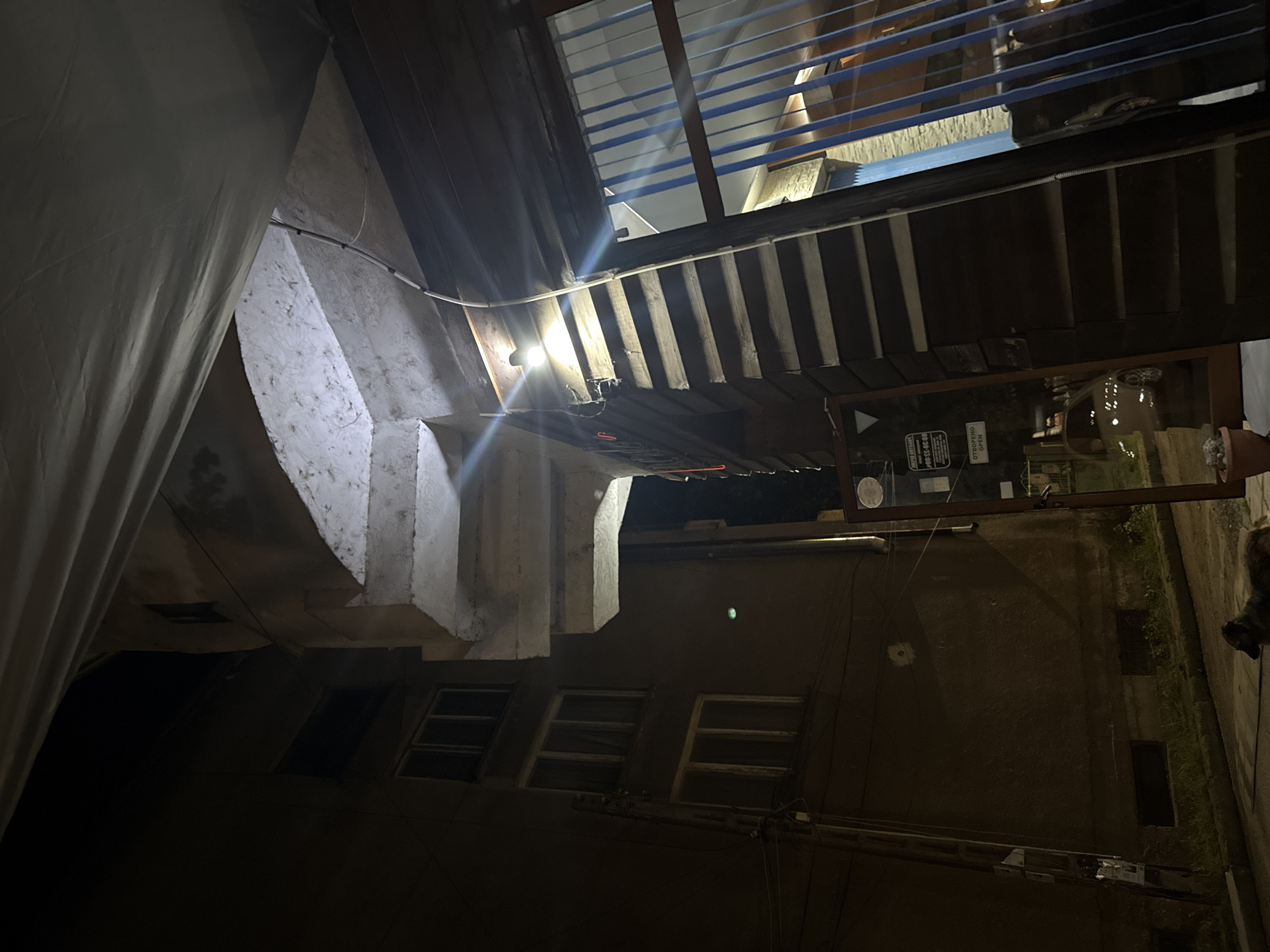 The image captures an outdoor or semi-outdoor space at night in Lom, Bulgaria, likely an alleyway, courtyard, or the side of a building complex. The scene is illuminated by artificial light sources, creating strong contrasts and shadows.
On the left, a multi-tiered concrete staircase or ramp ascends, featuring rough, textured surfaces. A large, dark curtain or tarp hangs down the far left side, partially obscuring the view. Running along the top edge of the concrete structure are exposed electrical wires within a conduit.
To the right, a wooden structure with horizontal planks and a ribbed, possibly corrugated metal or plastic awning/roof extends across the frame. A bright light fixture is attached to this wooden structure, emitting a strong glare and lens flare. Above this, blue-tinted horizontal blinds or slats are visible, suggesting windows or openings to an upper level.
Below the concrete staircase and the wooden structure, a building facade with multiple small, rectangular windows is visible. The wall is dark brown or grey. Numerous wires are strung across the scene, indicating electrical or utility connections.
Notable details include a large glass jar or container on a ledge below the wooden structure. On a sign below the wooden structure, the Cyrillic word "OTBOPEHO" (Open) is visible, along with its English translation "OPEN" and a phone number: "08-98-33-806". Another small sign with "ВХОД" (Entrance) is also partially visible. On the far right, near the ground, the head and shoulder of what appears to be a dog are present. The ground level shows a narrow strip of grass and a dark, possibly unpaved path. The overall impression is that of a functional, perhaps utilitarian, urban or semi-urban environment.