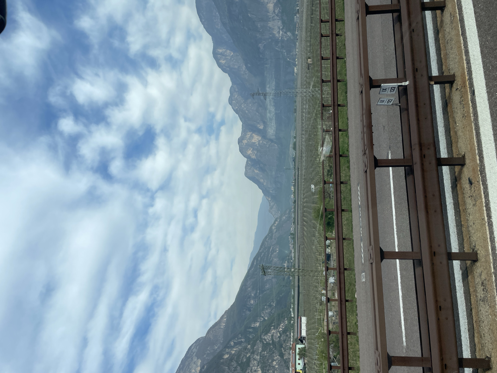 The image shows a mountainous landscape in Nave San Rocco, Italy. The view is from a roadside location, likely a highway or bridge, with a metal railing in the foreground. Beyond the railing is a stretch of cultivated land, possibly vineyards, with evenly spaced rows of plants. In the distance, large mountains rise, partially obscured by haze or clouds. Power lines and towers are visible, cutting across the landscape. The sky is blue with scattered white clouds. The road has a white line marking the lanes. The road appears to have mile markers labelled 122.