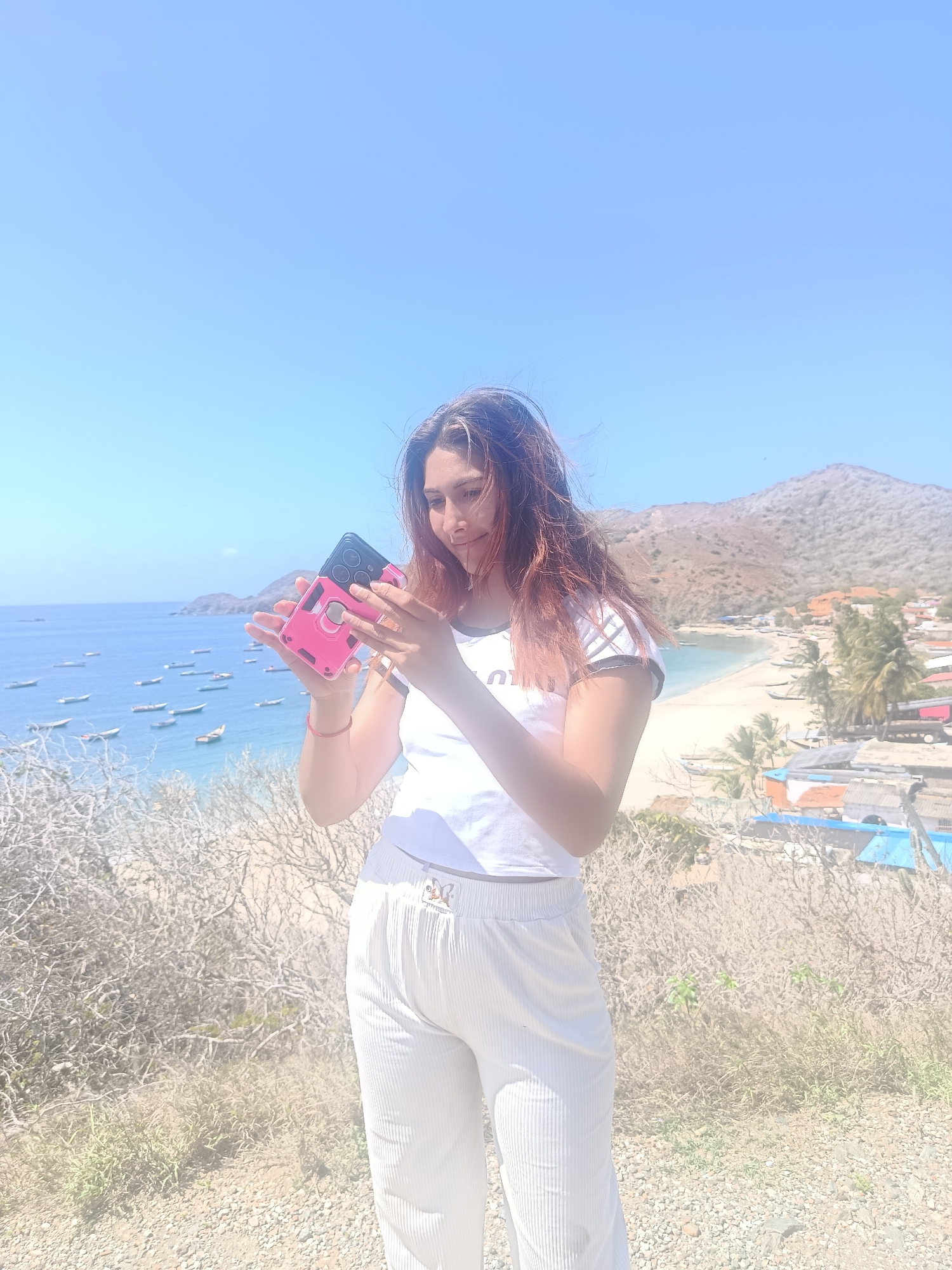 A young woman stands on a hill overlooking a bay in Manzanillo, Venezuela. She is wearing a white t-shirt and white striped pants, holding a pink phone case with a phone inside. The bay is filled with small boats, and a sandy beach with palm trees and buildings lines the shore. The background features a dry, mountainous landscape under a clear blue sky.