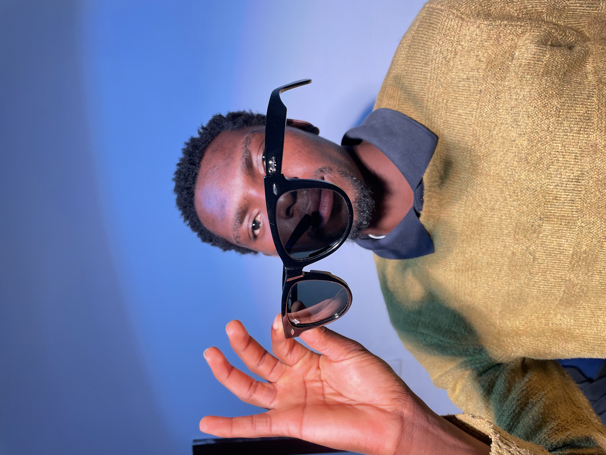 The image is a close-up of a man holding a pair of black sunglasses. The man is dark-skinned and has short, dark hair and a beard. He is wearing a dark-colored shirt and a tan sweater. The background is a blue gradient. The sunglasses are Wayfarer style. There is no information to suggest the photo was taken in Barakin Akawo, Nigeria.