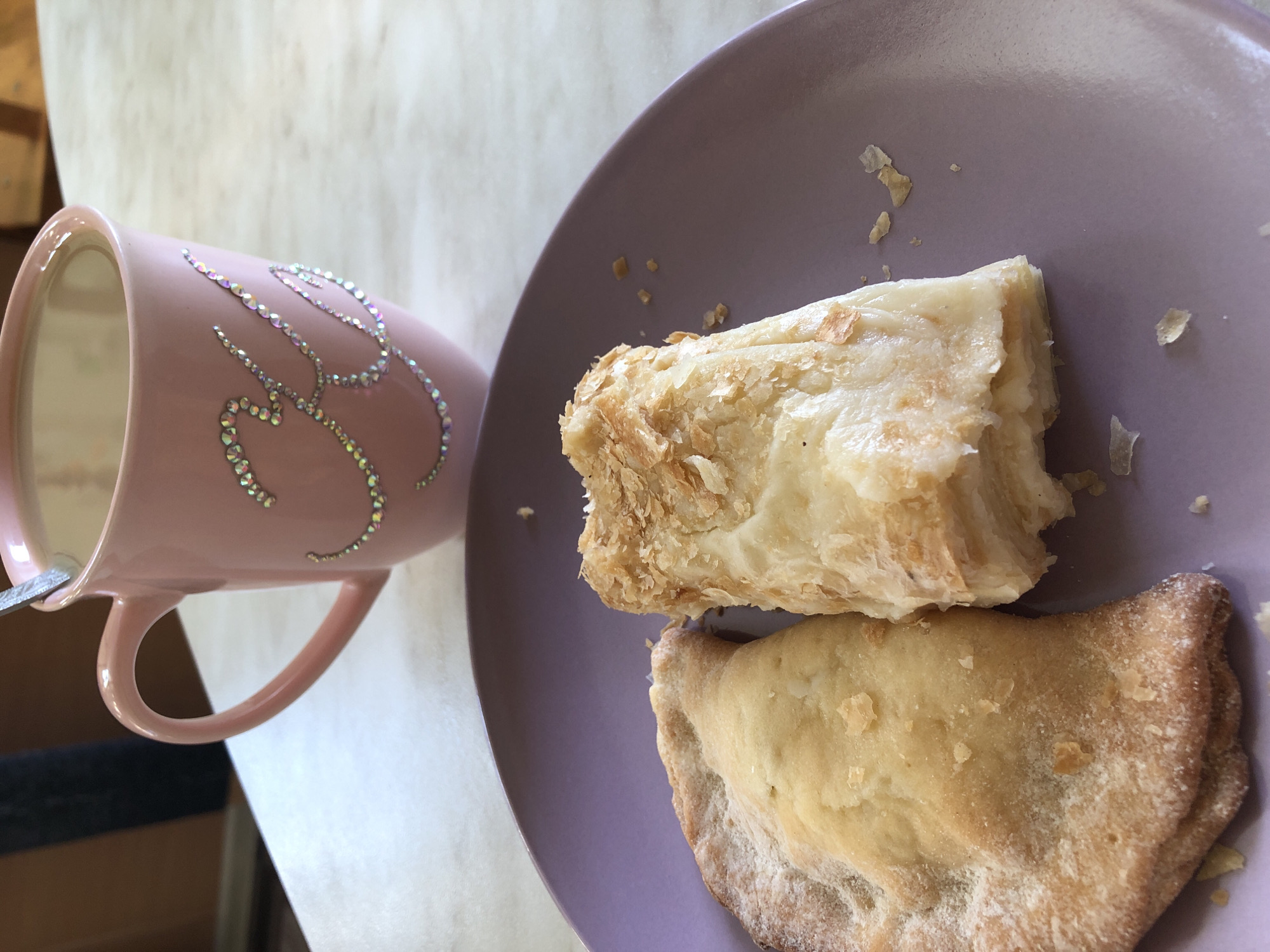 This image depicts a static indoor scene set on a light-colored, possibly marble or laminate, countertop. The location is contextualized as Noviy, Russia, though specific identifying architectural or geographical markers are absent.
In the upper portion of the frame, a pink ceramic mug with a handle is visible, containing a light-colored beverage, likely coffee or tea with milk. A metallic spoon is partially submerged in the liquid. The exterior of the mug is adorned with iridescent rhinestones forming what appears to be the capital letter 'K'.
In the foreground, a round, matte purple plate is positioned. On this plate, two distinct pastries are presented. To the left, a crescent-shaped pastry, possibly a turnover or hand pie, is visible. Its surface is a golden-brown hue and appears to be dusted with a white powder, likely powdered sugar. To the right of this, a rectangular pastry with a flaky, layered texture, consistent with puff pastry, is observed. Its exterior is golden-brown, and a lighter, possibly cream or custard, filling is visible between the layers. Small flakes of pastry are scattered on the purple plate around both items.
The background is softly blurred, showing a dark, undefined object, possibly furniture or clothing, against a lighter wall or surface. No individuals are visible in the frame, and no discernible actions are actively occurring. The lighting suggests ambient or natural light.