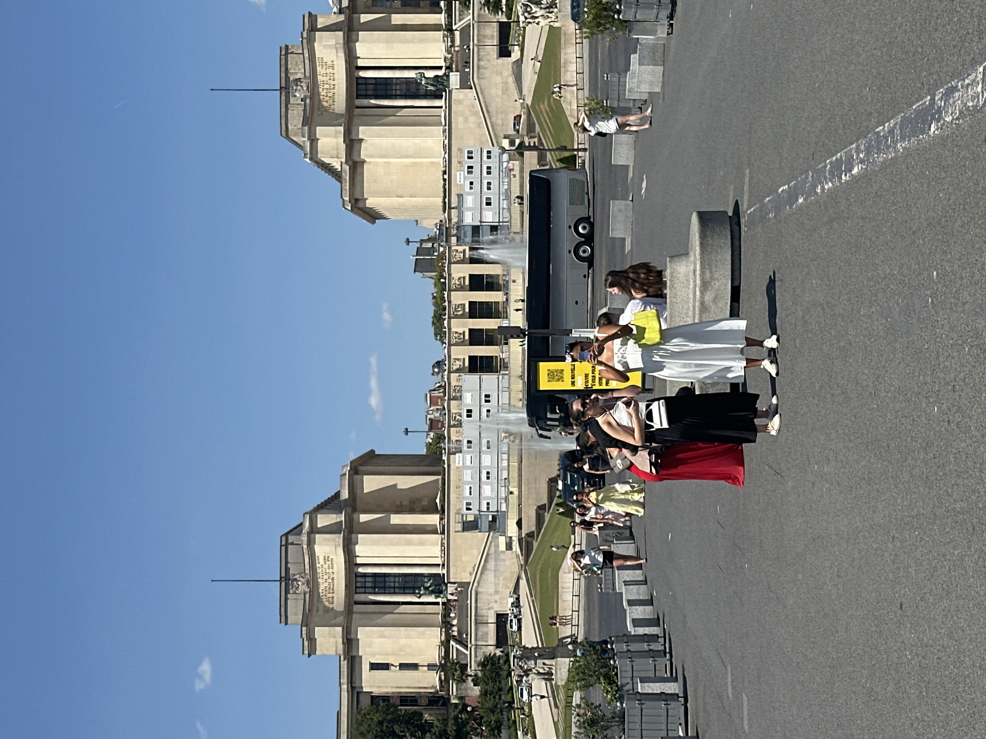 The image depicts a wide-angle, eye-level view of the Palais de Chaillot in Paris, France, under a clear blue sky. In the foreground, a paved area with several people walking and standing is visible. Two women in the center are holding cell phones, one appearing to take a picture. One woman wears a white dress and the other a black skirt and white top. Further to the left, a woman in a long red dress stands with two other individuals. A silver tour bus is parked in the middle ground, with a bright yellow advertisement attached to its side. Behind the bus, a large fountain sprays water. The Palais de Chaillot, a grand neoclassical building with a semi-circular facade and two wings, dominates the background. Its stone exterior is adorned with inscriptions and statues. Terraced gardens with greenery and pathways lead up to the building. The overall scene suggests a sunny day in a popular tourist area.