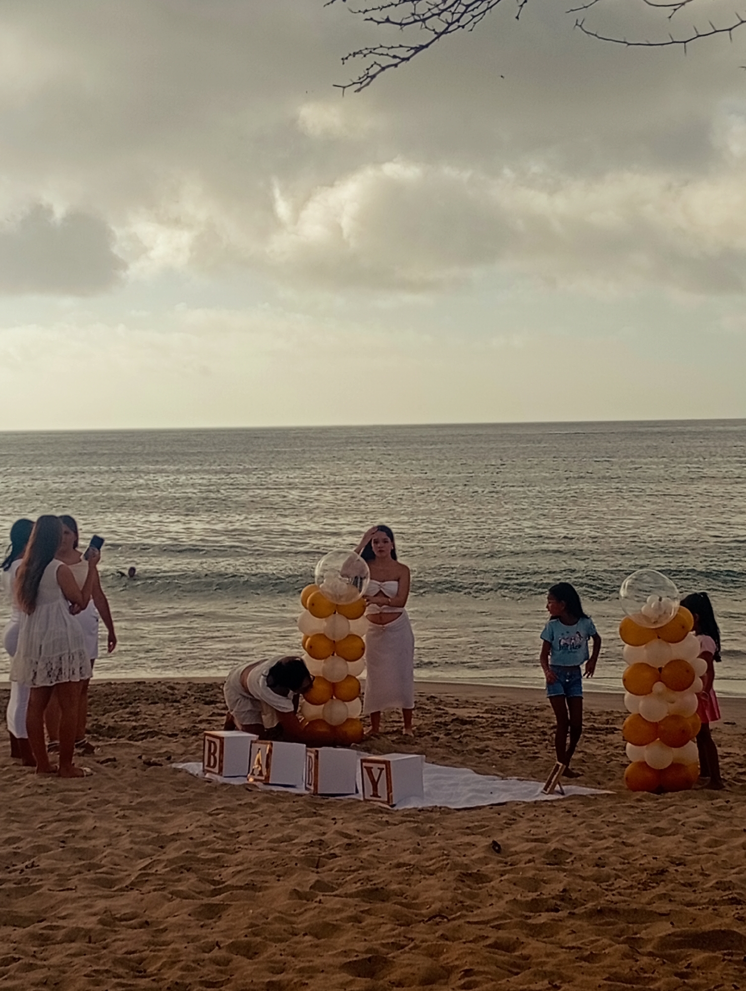 The image depicts a beach scene, likely a baby shower celebration, in Pedro González, Venezuela. A group of people, mostly women dressed in white, are gathered on the sand. A pregnant woman, also in white, stands prominently. Two balloon columns, colored yellow and white, flank a "BABY" arrangement made of block letters placed on a white cloth. One woman is kneeling near the block letters, apparently adjusting or arranging them. Another woman holds a phone, possibly taking pictures. The ocean stretches to the horizon under a cloudy sky. In the background, a person is seen swimming in the water. A branch with bare twigs is visible in the upper part of the image.