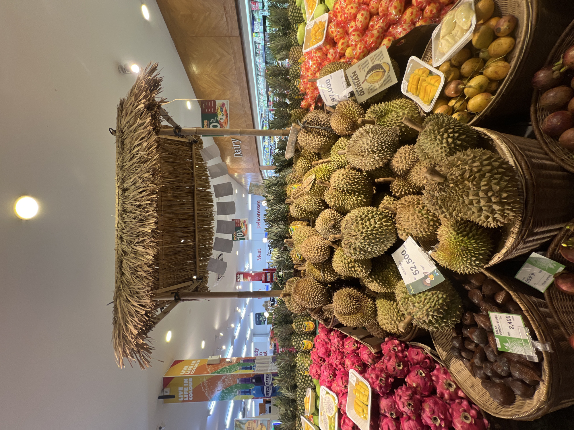 This image captures a vibrant fruit display within what appears to be a supermarket or grocery store in North Kuta, Indonesia. The central focus is a large, inviting presentation of various tropical fruits, predominantly durians piled high in traditional wicker baskets, some under a rustic thatched roof structure.
Other abundant fruits include bright pink dragon fruits, dark brown salak (snake fruit), and green pineapples. Nearby baskets contain whole mangoes and passion fruits, with some mangoes also presented pre-cut in clear plastic containers. The setting indicates an indoor retail environment, characterized by bright overhead lighting and visible supermarket sections in the background, labeled "Dairy," "Meat," and "Delicatessen." A large banner for "BREEZER BREEZER" beverages is also visible, with the slogan "LIVE LIFE IN COLOUR."
The scene is designed to attract shoppers, showcasing fresh produce. Price tags are prominently displayed on many items, providing specific details. For instance, a sign next to the durians reads "DURIAN LOKAL 52,500" (Local Durian, priced at 52,500, likely Indonesian Rupiah), and another large sign advertises "EMPAT MANFAAT DURIAN" (Four Benefits of Durian). Another price tag shows "97,000" for durian. Salak Pondoh Super is priced at "2,400," and another item is marked "4,450." A "DIS 10%" (Discount 10%) sign is also seen. The abundance and variety of tropical fruits suggest a lively market or grocery experience.