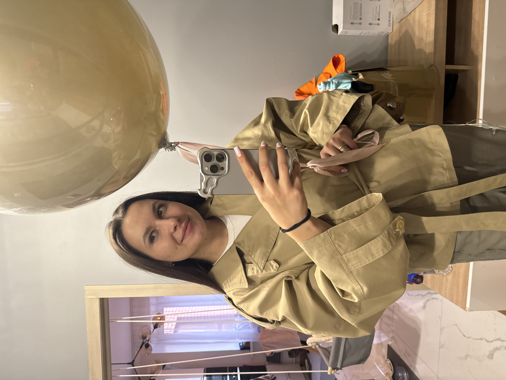 A woman is taking a selfie in an interior space. She is holding up her phone, which is decorated with a silver case. Attached to the phone is a large, opaque gold-colored balloon with a pink ribbon. The woman is wearing a tan trench coat over a white shirt. A black wristband adorns her left wrist. She has long dark hair and is looking at the camera with a slight smile.
Behind the woman, there's a glimpse of a room with what appears to be a clothes drying rack near a window with horizontal blinds. To the upper right, some clothing items (orange, blue) are on top of a wooden dresser or storage unit. Based on the visible interior style and the person's attire, the location appears to be indoors, possibly in Odintsovo, Russia.