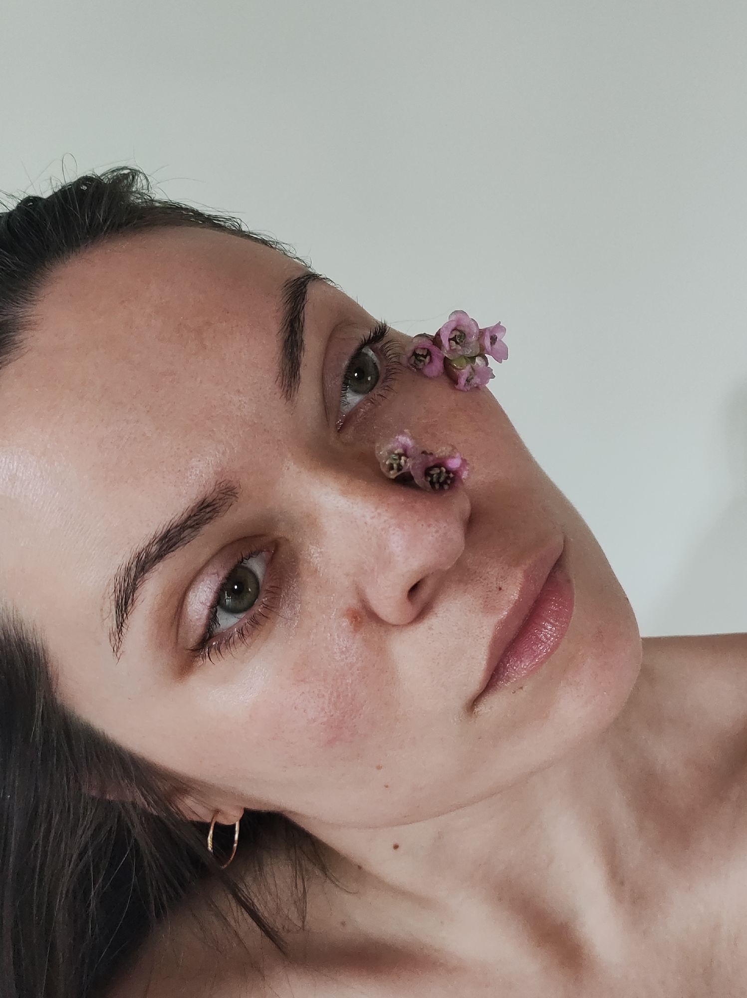 A close-up, indoor shot presents a light-skinned woman with flowers placed on her face. The woman has dark hair pulled back, revealing a gold hoop earring in her left ear. Her eyes are green, and her eyebrows are dark and defined. Small pink flowers are positioned near her left eye and on the bridge of her nose. Her lips are slightly parted and appear to have a glossy sheen. The background is a plain, off-white color. The location and city are unknown.