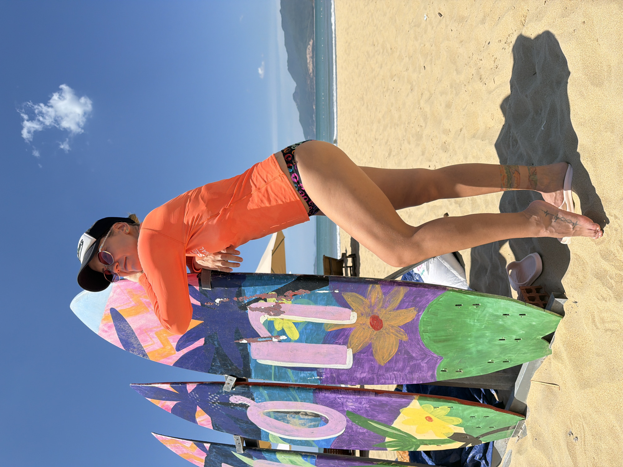 A woman is standing on a sandy beach in Quy Nhon Ward, Vietnam, with three surfboards to her left. She is leaning against one of the surfboards, with her right arm resting on it. The surfboards are colorful, with painted designs. The woman is wearing an orange long-sleeved rash guard and black and multi-colored patterned bikini bottoms. She has sunglasses and a black and white baseball cap on. The sky is blue with a single white cloud. In the background, the ocean meets the horizon with a line of mountains in the distance. There are beach chairs and an umbrella visible behind the surfboards. The sand is light brown and there are shadows cast on it.
