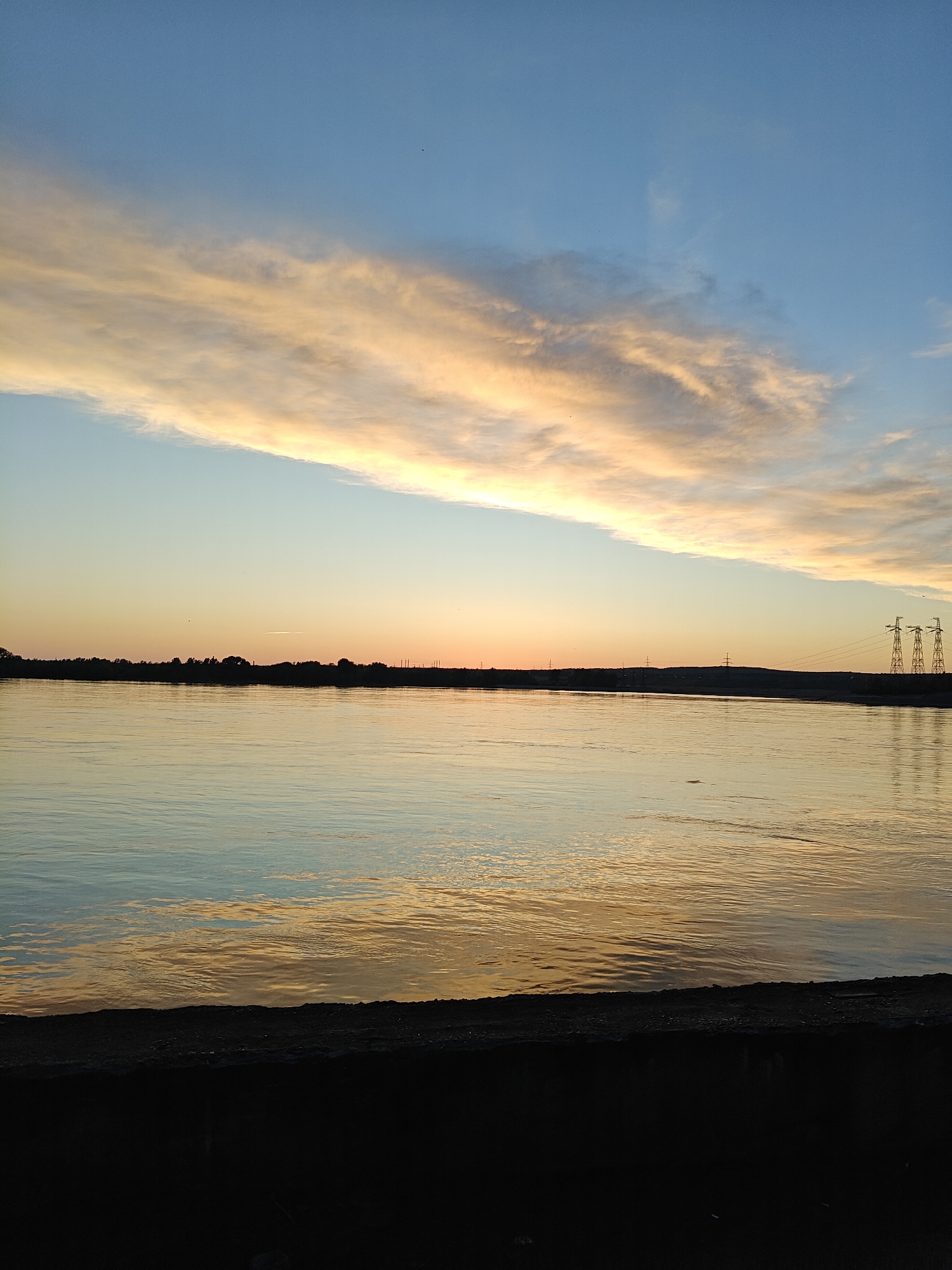 This media file presents a wide landscape view of a calm body of water, likely a river or large reservoir, captured during either sunrise or sunset, given the warm, low light. The scene is tranquil and devoid of people.
The upper portion of the sky is a clear, serene blue, gradually transitioning downwards into a band of soft, elongated clouds streaked with warm golden, orange, and pale yellow hues. These clouds extend horizontally across the middle of the sky, catching the light from the setting or rising sun. A few tiny, dark specks, possibly distant birds, are barely visible against the vast sky.
The expansive water surface beautifully reflects the sky's colors, showing gentle ripples that distort the reflections into shimmering patterns of blues, golds, and oranges. The distant horizon features a dark, silhouetted shoreline, heavily treed, which stretches across the image. On the right side of this distant land, several large power line pylons are distinctly visible as dark, angular silhouettes against the brighter sky.
In the foreground, a dark, rough-textured embankment or wall occupies the bottom portion of the frame, providing a strong base to the composition. This structure appears to be made of concrete or stone, suggesting a man-made edge to the waterway.
The overall impression is one of peaceful natural beauty, with the warm light painting the sky and water in rich, contrasting colors. There is no visible text in the image. Given the location context of Chaikovsky, Russia, this scene likely depicts the Kama River or Votkinsk Reservoir.