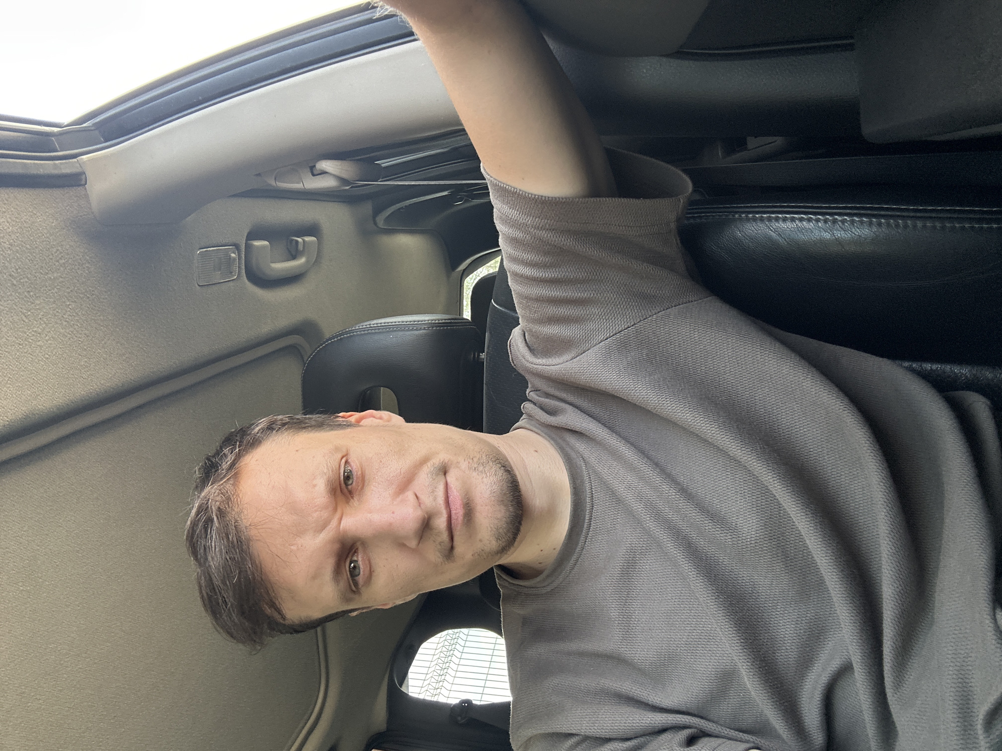 A selfie taken from inside a vehicle shows a man with short brown hair and a light beard. He is wearing a grey t-shirt and looking at the camera with a slightly furrowed brow and a neutral expression. The man is seated in the driver's seat of the car, with the steering wheel and dashboard visible in the background. Sunlight is coming in from the right side of the frame, illuminating his arm and the interior of the car. The exterior of the car shows a wire fence and some greenery through the window. The vehicle appears to be parked, possibly in an outdoor location with a fence. There are no other people or animals present in the image. The location is Shatava, Ukraine.