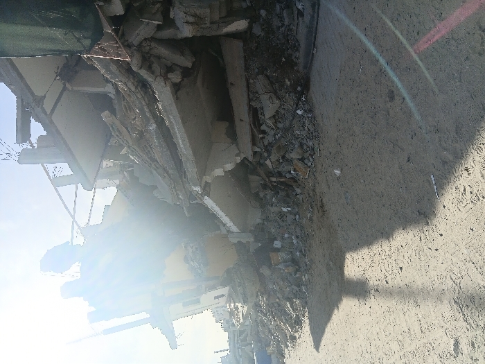 The image depicts the aftermath of severe structural damage to a multi-story concrete building in Khan Yunis, Palestinian Territory, under clear, bright daylight. The building is largely collapsed, with multiple floors reduced to extensive piles of concrete rubble, twisted rebar, and shattered wall fragments. Exposed interior sections reveal portions of a yellow-painted wall. The foreground consists of light brown, sandy ground showing numerous footprints. A prominent, dark shadow of an unseen object extends diagonally across the sandy foreground from the left. Intense sun glare from the upper left portion of the frame overexposes part of the damaged structure and produces a lens flare. A dark green netting or tarp is visible along the far right edge of the image, partially obscuring the wreckage. No individuals are present in the scene.
