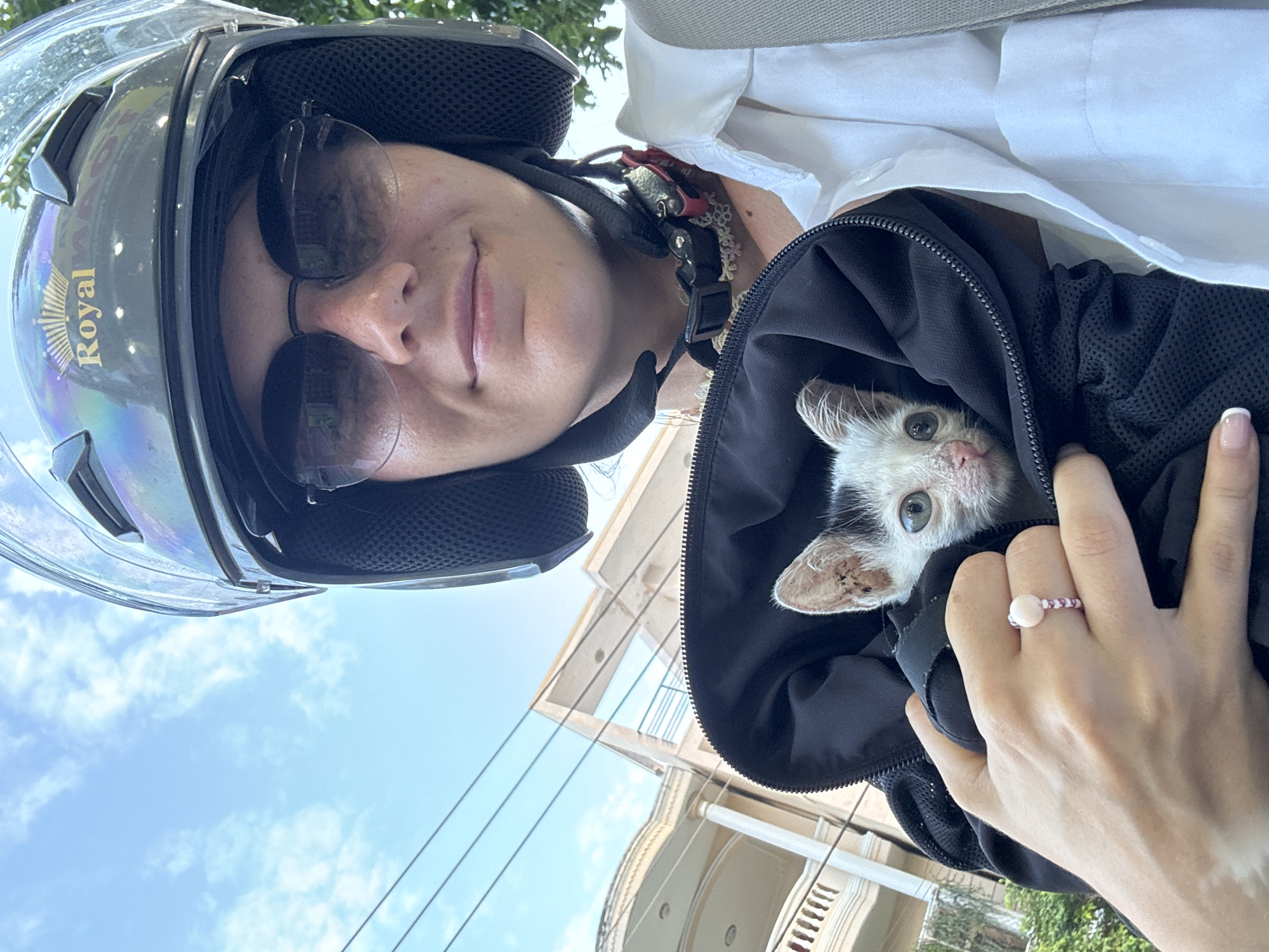 A woman wearing a black motorcycle helmet with a "Royal" logo and sunglasses is taking a selfie. She is holding a very small, skinny kitten with white fur and black markings inside a black bag. The kitten appears to be distressed and is looking towards the camera. The woman is wearing a white collared shirt. In the background, there is a blue sky with some clouds, utility lines, and a building with columns, suggestive of a residential area. The context provided indicates this scene is in Phan Thiết, Vietnam.