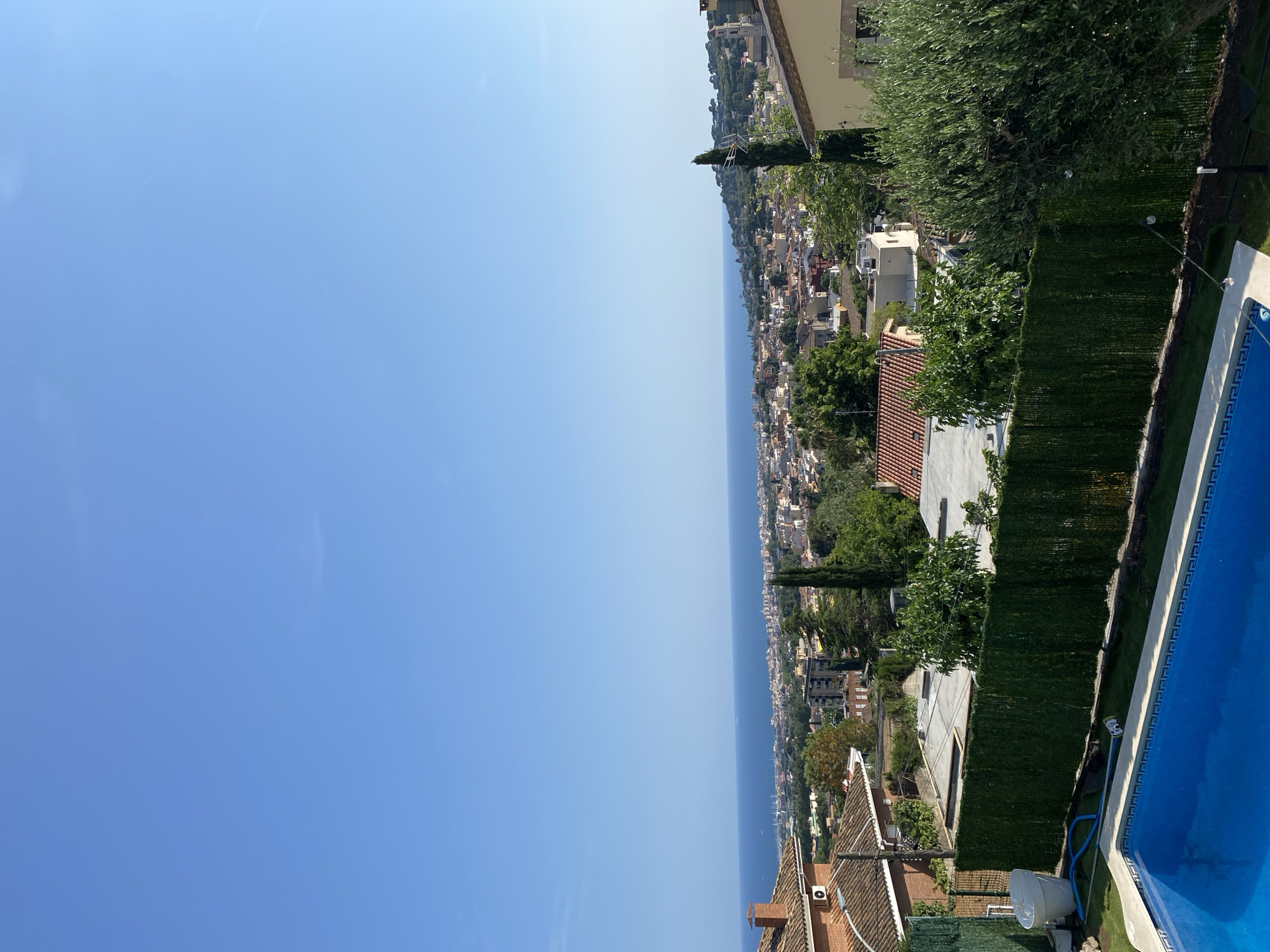 The image is a landscape scene taken from a high vantage point in what appears to be the residential area of Vilassar De Dalt, Spain. A clear blue sky occupies the majority of the left half of the frame. The right half of the image captures the urban sprawl leading towards the coastline, where the sea meets the land. The scene includes a swimming pool in the foreground. Houses with terracotta roofs dot the landscape, interspersed with green trees and shrubs. A tall, slender cypress tree stands out among the greenery. The overall impression is one of a Mediterranean landscape with a combination of residential buildings and natural elements.
