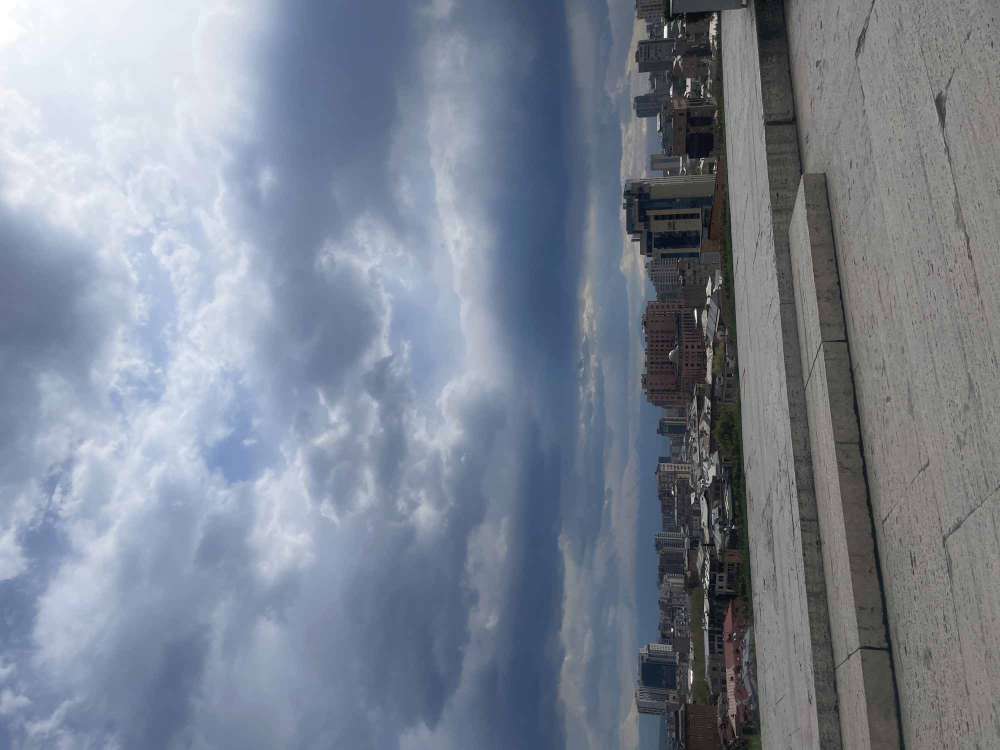 The image captures an expansive urban landscape beneath a dramatic and heavily clouded sky, viewed from an elevated perspective. The sky occupies a significant portion of the scene, featuring large, textured clouds that range in color from bright white, illuminated by patches of sunlight, to dark, brooding grey, suggesting dynamic weather conditions. Sections of clear blue sky are visible intermittently through the cloud cover.
The mid-ground and background are dominated by a dense cityscape. Numerous buildings, varying in height, age, and architectural style, stretch across the horizon. Some structures appear modern with glass facades, while others are older, built from stone or brick. The urban development extends far into the distance, giving way to a hazy horizon under the overcast sky. Green foliage can be seen interspersed among the buildings, indicating trees or park areas within the city.
In the right foreground, a substantial, light-colored stone or concrete structure with visible horizontal layers or steps frames the view. This textured element, possibly part of a grand staircase, monument, or large building, indicates the high vantage point from which the photograph was taken.
The scene depicts daytime, likely late morning or afternoon, characterized by the natural light, despite the heavy cloud cover. The weather appears unsettled or transitional, with the striking cloud formations. No people or specific activities are visible within the frame. There is no visible text. The location context for this urban view is Yerevan, Armenia, with the foreground structure possibly being part of a landmark like the Cascade Complex.