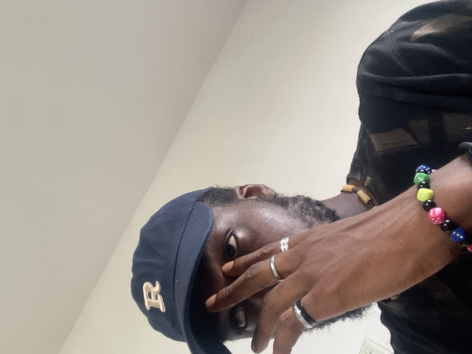 The image shows a close-up of a person wearing a dark blue baseball cap with an embroidered 'R' on the front. The person is a Black male with a beard and is wearing several rings on his left hand, including a silver band, a set of three stacked silver bands, and a black and silver ring. He is also wearing a beaded bracelet with dice-shaped charms in various colors (pink, blue, green, yellow) and a necklace made of wooden beads. The person is holding his hand up to his face, obscuring part of his eyes. He is wearing a dark t-shirt that appears to have a bleached or tie-dye pattern. The background is a plain, light-colored wall, possibly a ceiling, with a subtle diagonal line indicating a corner. The lighting suggests an indoor environment. There are no identifiable landmarks or contextual clues in the image that would indicate a specific location in Abuja, Nigeria.