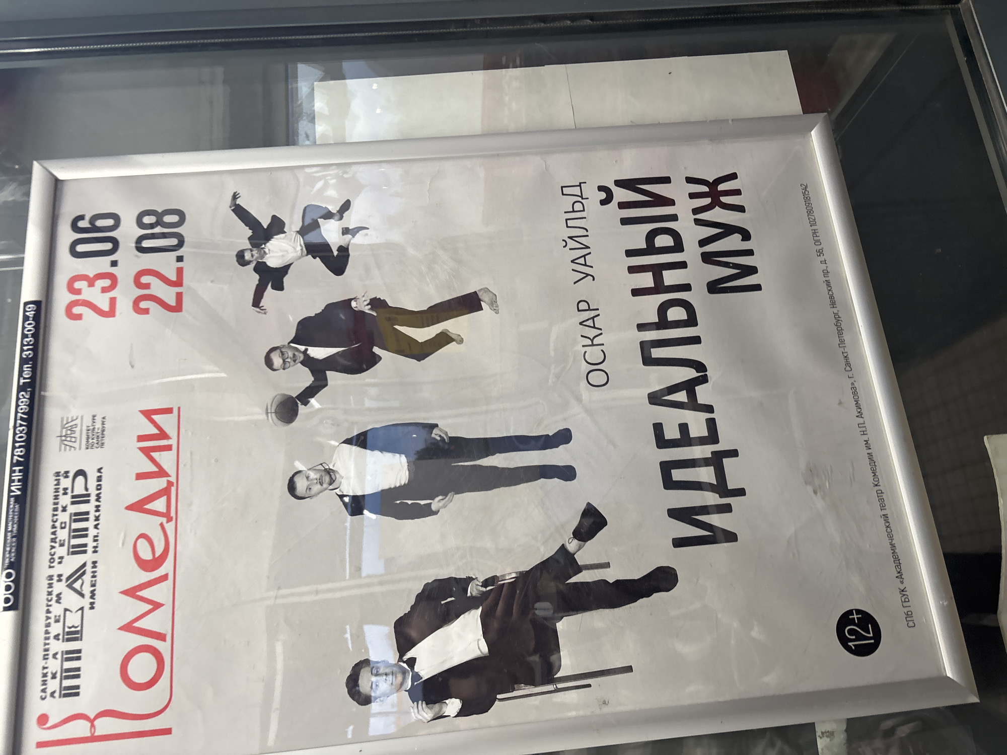 The image shows a poster for a play titled "The Ideal Husband" by Oscar Wilde, displayed in a glass-covered frame. The poster features several stylized images of men in formal attire, some appearing to be in mid-action, possibly representing characters from the play. The text on the poster is in Russian and includes the name of the theater, "АКАДЕМИЧЕСКИЙ ТЕАТР КОМЕДИИ ИМЕНИ Н.П. АКИМОВА," and performance dates "23.06" and "22.08." There is also a "12+" age rating symbol. The poster is illuminated, and reflections are visible on the glass, indicating it is likely displayed in a public space, such as a theater lobby or a street-facing window. The background appears to be a blurred interior or exterior scene. The poster itself is framed and mounted on a wall or in a window.