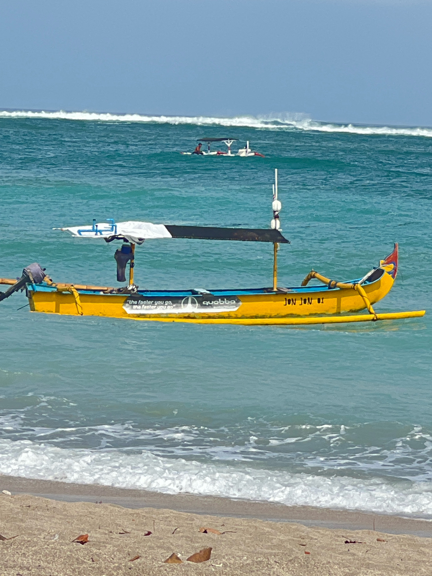 A bright yellow traditional Balinese outrigger canoe, named "JON JON 01", floats on turquoise waters off the coast of Kuta, Indonesia. The canoe is equipped with a small outboard motor at the stern and an awning providing shade over the center. The words "the faster you go, the faster you go" are visible on the side of the boat, accompanied by a circular logo. In the background, several other boats are visible on the ocean, with large waves breaking further out. The shoreline is a sandy beach with sparse dry leaves scattered across it. The sky is a clear, pale blue.