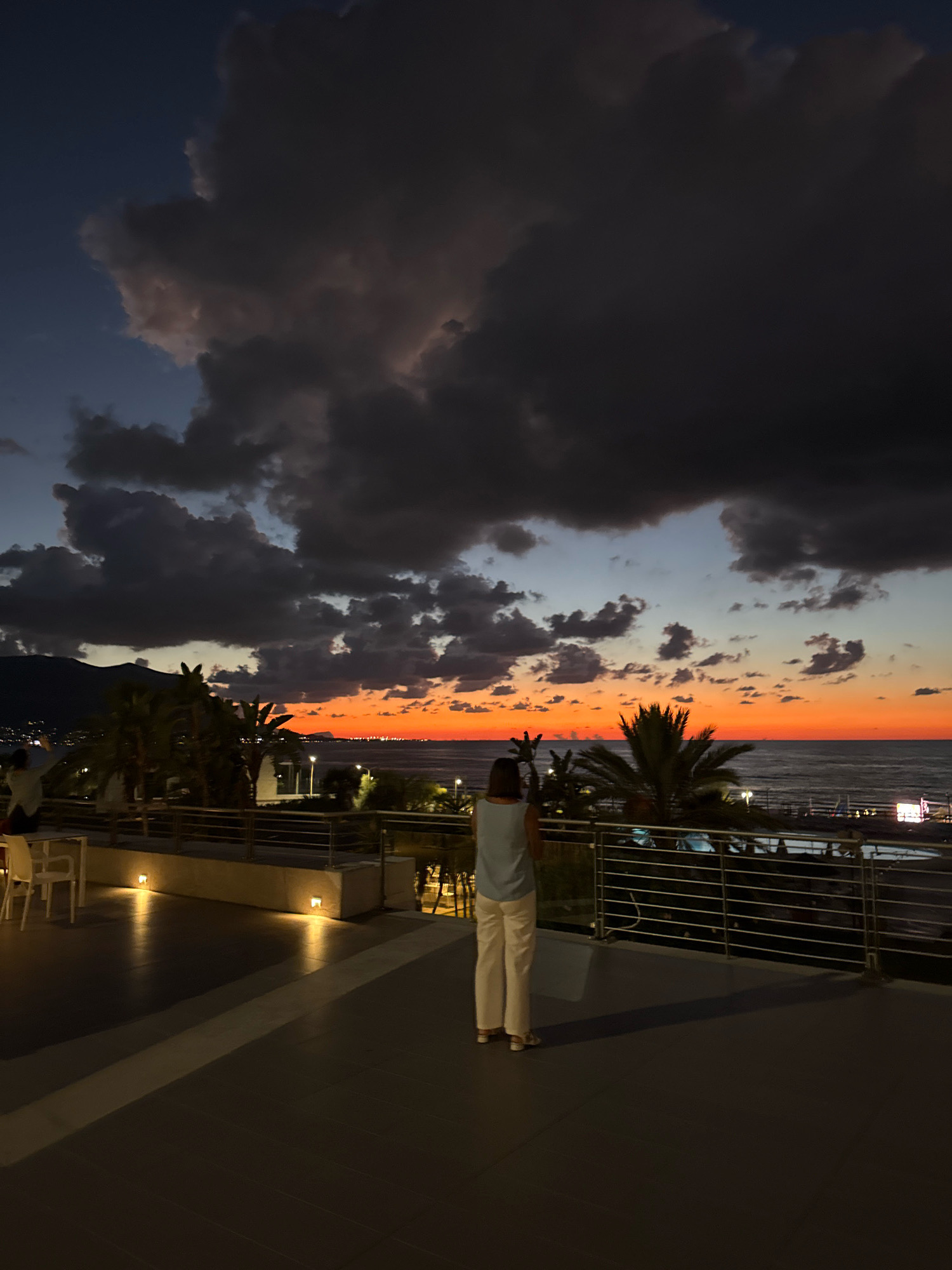 A woman stands on a balcony overlooking a beach at dusk in Isola delle Femmine, Italy. She is wearing a light blue top and white pants. The sky is a dramatic mix of dark clouds and a vibrant orange sunset over the sea. Palm trees and some buildings are visible along the coastline. The balcony is paved with dark tiles, and a metal railing runs along the edge. There are also some tables and chairs on the balcony, suggesting it is part of a restaurant or hotel. The lighting is dim, indicating it is shortly after sunset.