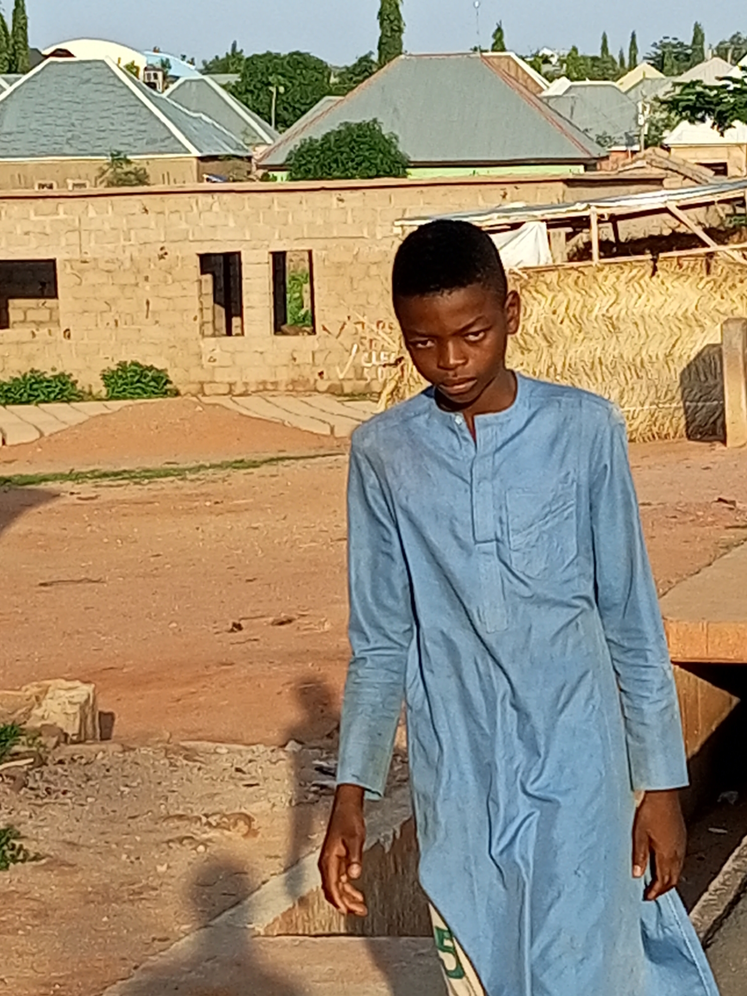 A young boy with dark skin and short black hair stands in the foreground, looking towards the viewer with a neutral expression. He is wearing a light blue, long-sleeved garment with a Mandarin collar and a button placket. The garment appears to be made of cotton. The boy is positioned in a dirt courtyard with sparse vegetation and some debris. Behind him, a wall constructed from light-colored bricks is visible, with several openings that appear to be windows or doorways. Beyond the wall, there are buildings with sloped roofs, some covered in gray shingles. The overall environment suggests a residential area in a warm climate, consistent with Pantami, Nigeria. The lighting indicates it is daytime, likely late afternoon given the shadows.