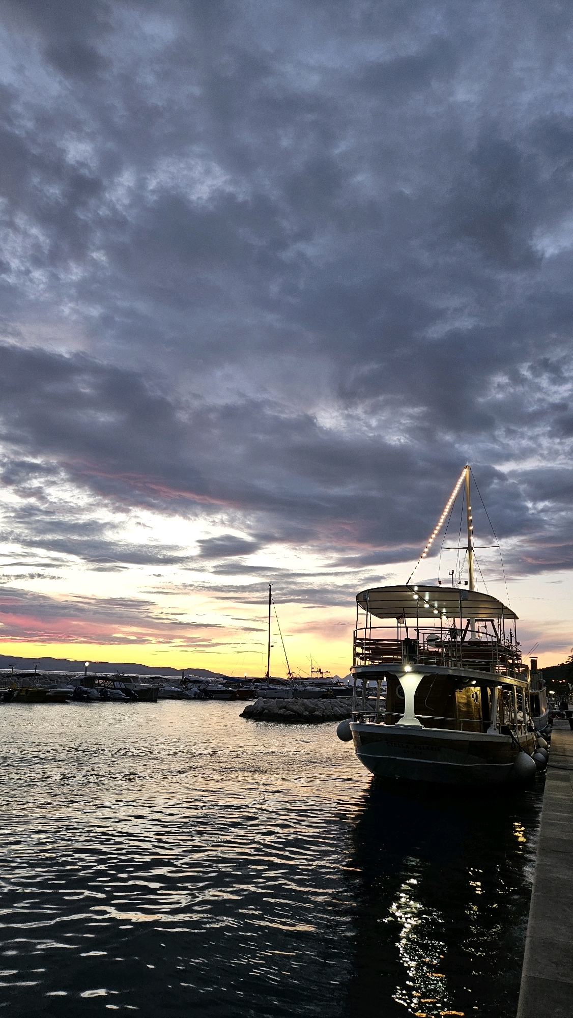 A docked boat with lights strung along its mast is anchored in a harbor at dusk. The boat is positioned on the right side of the frame, facing left, with its reflection shimmering on the water. In the background, a row of smaller boats and a distant shoreline are visible under a dramatic sky filled with dark, heavy clouds tinged with pink and orange hues from the setting sun. The water is calm, with gentle ripples reflecting the light from the boat and the sky. The scene is likely in Općina Tučepi, Croatia, known for its picturesque harbors and coastal beauty.