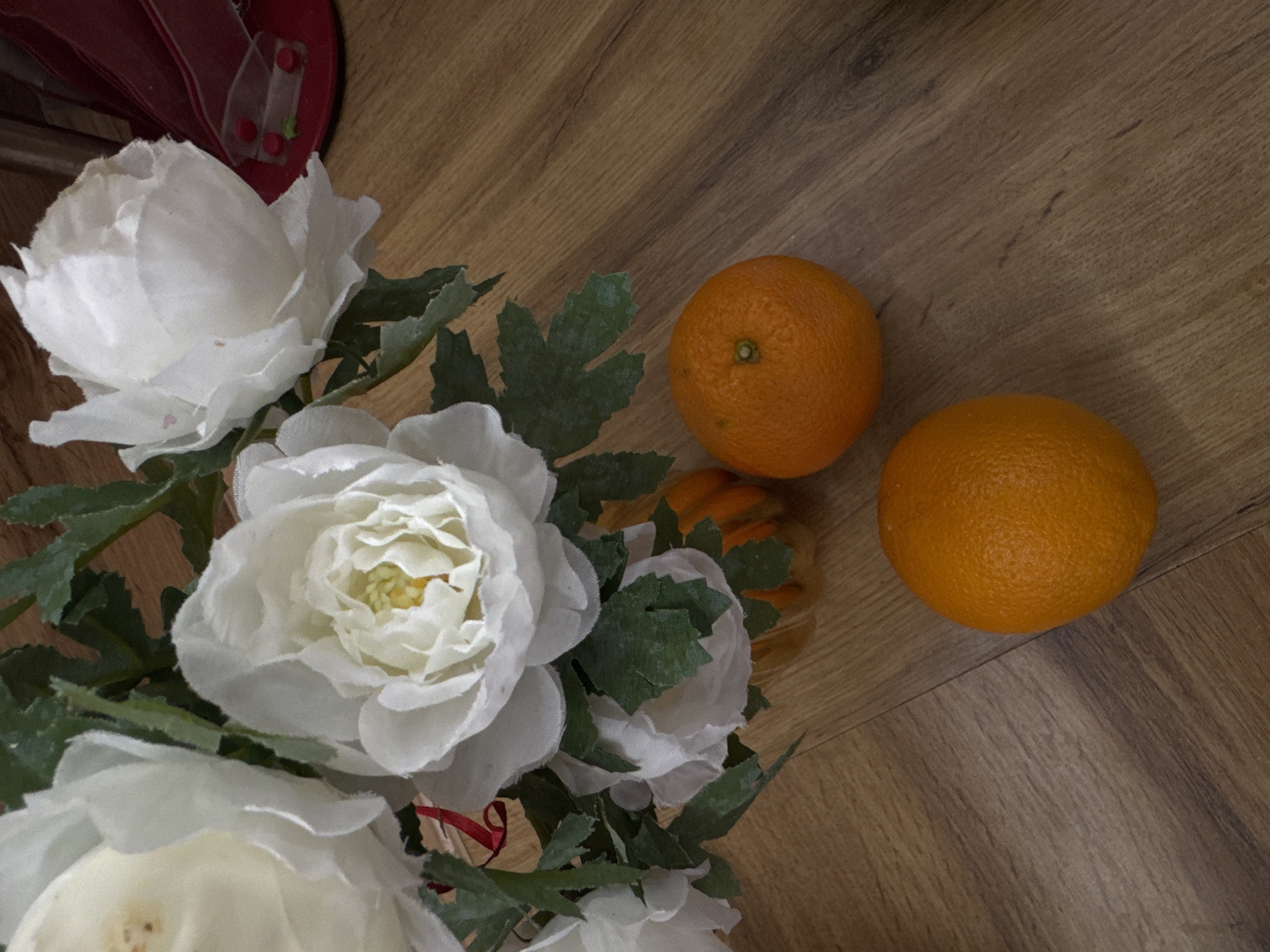 The image depicts a still life with two oranges and a bouquet of white flowers on a wooden surface. The oranges are positioned to the right of the flowers. A glimpse of a red object is visible in the upper left corner. The environment appears to be indoors, suggested by the wooden surface and the composition of the objects. There is no evidence of the location being Chaikovsky, Russia.
