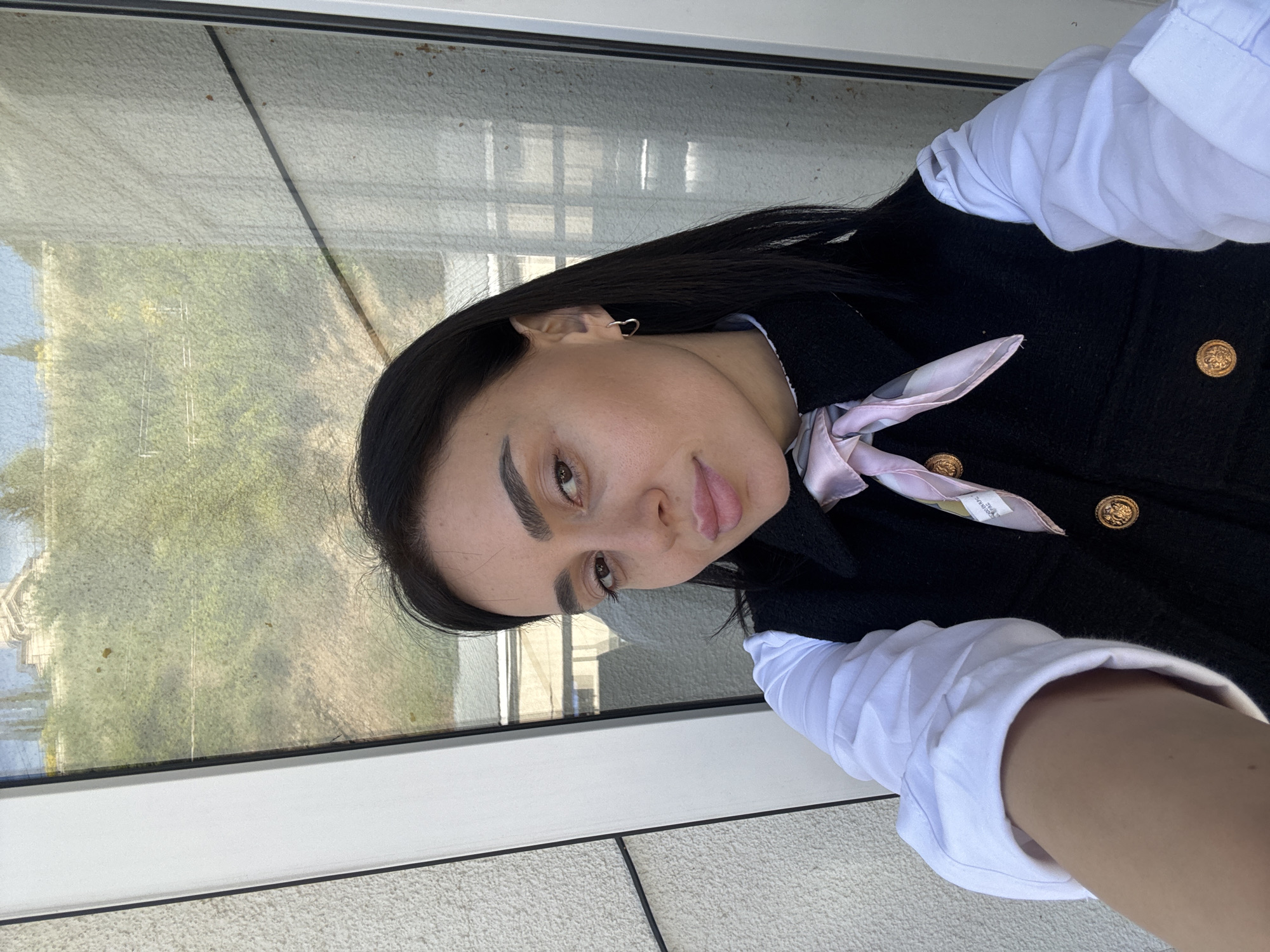 A young woman is captured in a selfie-style photograph, rotated 90 degrees counter-clockwise. She has long, dark, straight hair, prominent eyebrows, dark eyes, and full lips, looking directly at the camera with a calm, poised expression. She is wearing a white collared shirt underneath a black jacket or vest, which features ornate, gold-toned buttons. A light pink and white patterned scarf is tied neatly around her neck, and a small, simple hoop earring adorns her left ear.
The setting appears to be indoors, positioned next to a large window that offers a view of the outside. Through the window, green trees and what seem to be distant buildings or structures are visible, indicating an outdoor scene during clear daytime conditions. The window glass itself has some texture or light grime, with reflections of the interior. Below the window, a clean white frame and a textured light-colored wall or sill are visible. A small, blurry label with faint, unreadable text is visible on the scarf. The overall impression is a casual portrait taken in a well-lit indoor space overlooking a vibrant exterior, consistent with the provided location context of Tbilisi, Georgia.