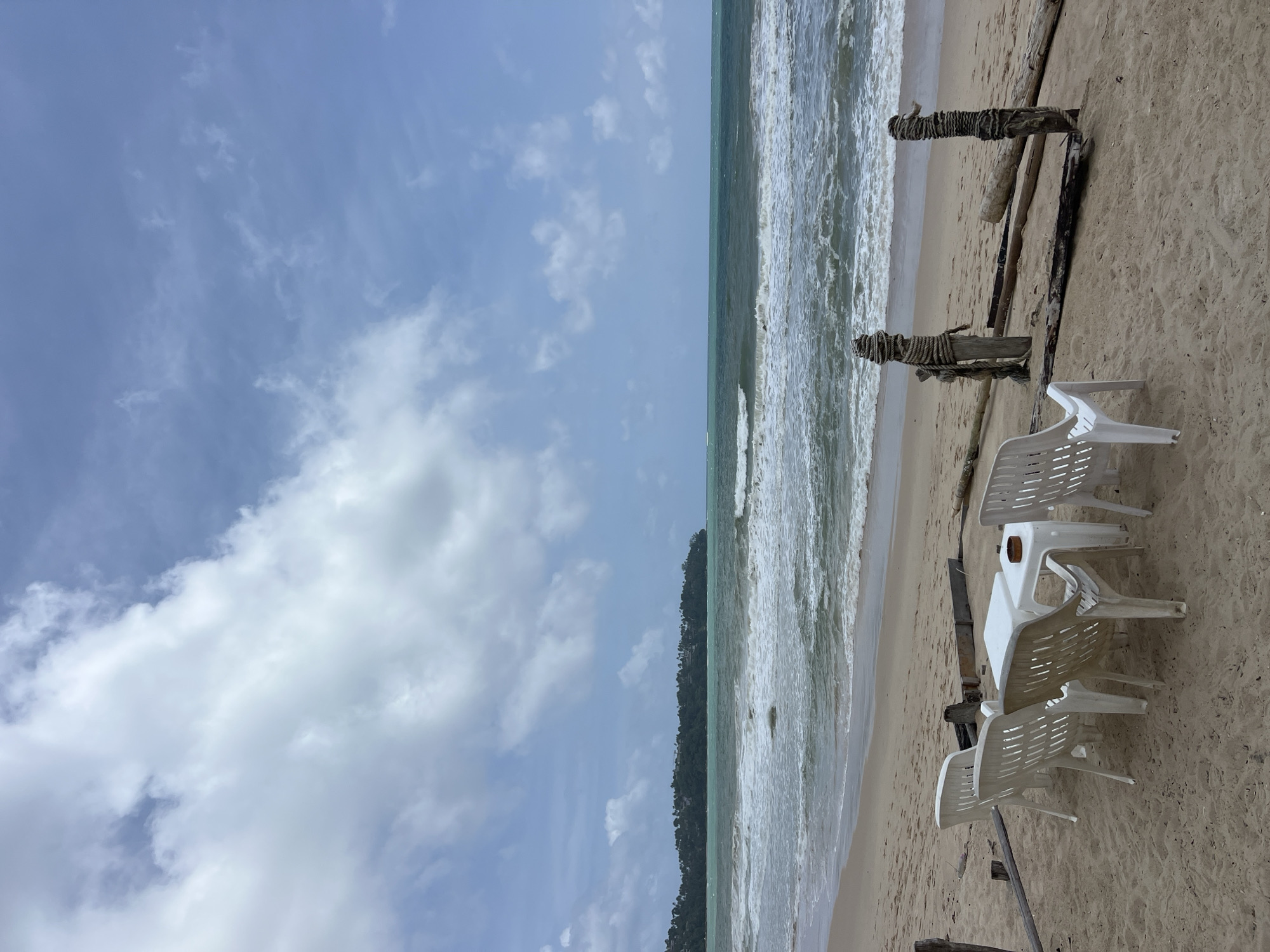 This is a photograph of a beach scene in Choeng Thale, Thailand. The foreground features a sandy area with four white plastic chairs arranged around two white plastic tables. On one of the tables, a small, circular, brown object, possibly an ashtray with a stubbed-out cigarette, is visible. Two weathered wooden posts, wrapped with rope, stand behind the chairs and tables. Beyond the sandy area, the ocean stretches to the horizon with moderate waves breaking on the shore. A distant treeline with some structures can be seen on a headland to the left, and the sky above is partly cloudy with patches of blue.