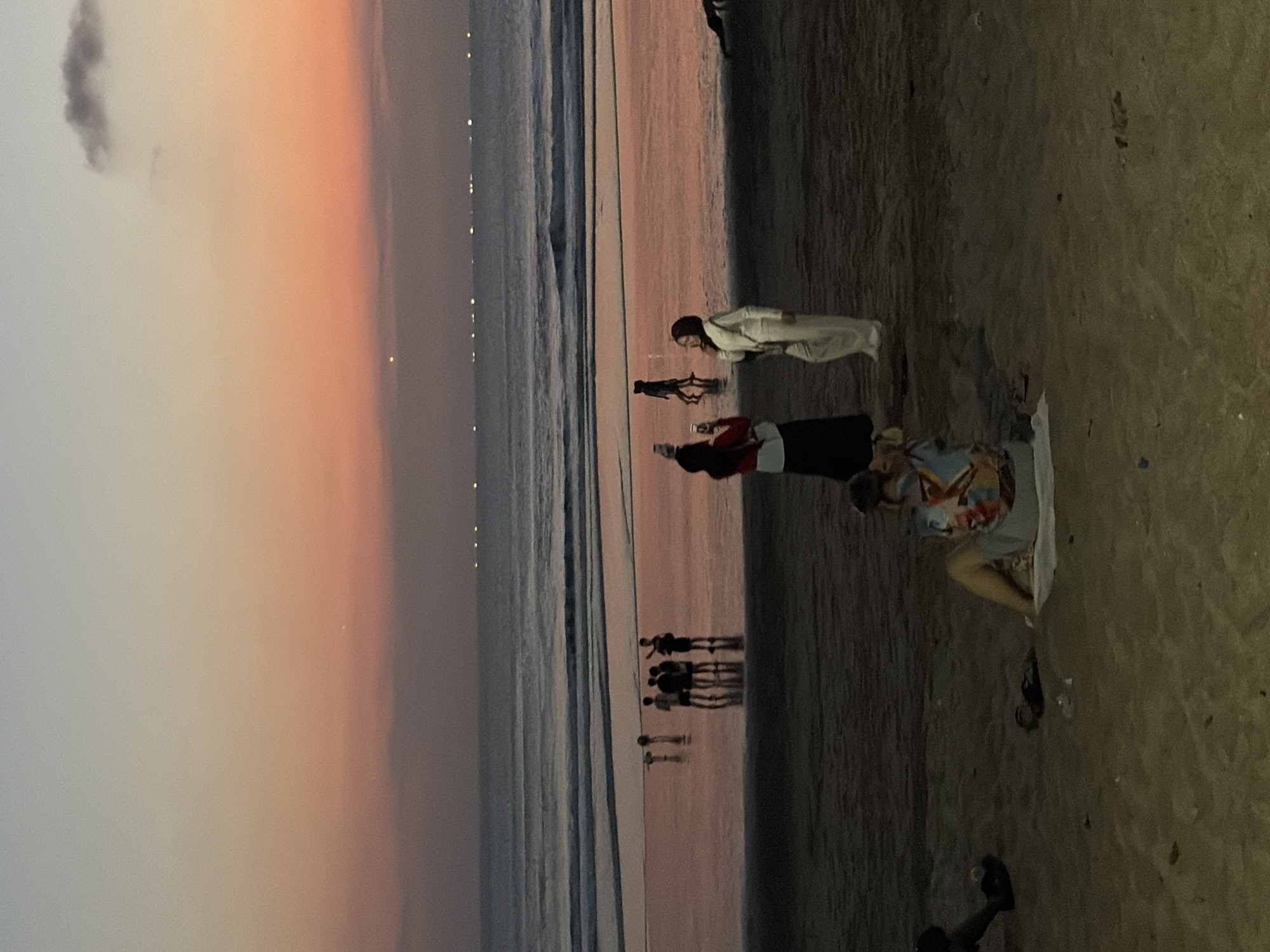 At sunset on Kuta Beach, Indonesia, several people are visible on the sand and in the shallow ocean. In the foreground, a person sits on the beach with their back to the camera, wearing a colorful shirt and gray shorts. Further back, near the water's edge, two individuals stand together; one woman in white appears to be taking a photo with her phone, while another woman in a red top and black skirt looks on. A group of four individuals, likely young men, stand closer to the ocean, their reflections visible in the wet sand. In the distance, a line of lights stretches across the horizon, indicating activity on the water. The sky displays a gradient of colors, transitioning from a pale blue at the top to warm orange and pink hues near the horizon, characteristic of a tropical sunset. The waves are gentle, creating a calm atmosphere on the beach.