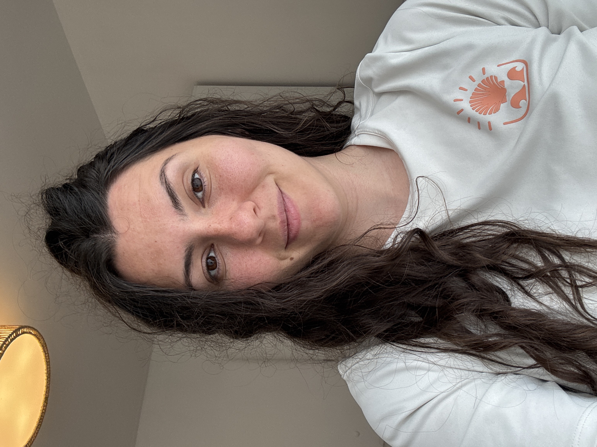 A person with brown hair and light skin is wearing a white long-sleeved shirt with an orange seashell logo on the right side of the shirt. The person is looking directly at the camera and smiling slightly. A lamp is visible in the bottom left corner of the image. The background appears to be a plain wall. The location is unknown.