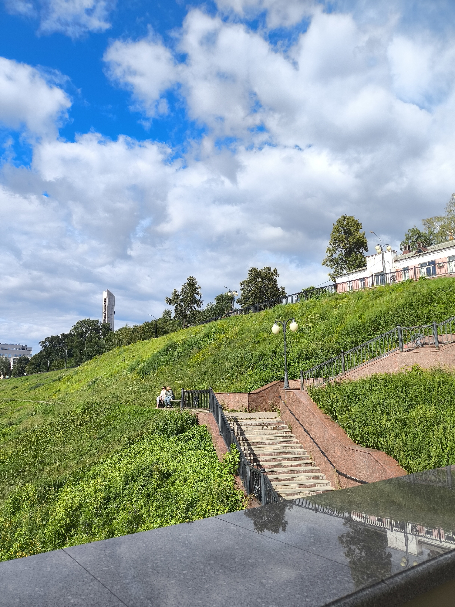 The image depicts a view of the Mikhail Petrovich D'Agostino embankment in Izhevsk, Russia, on a cloudy day. In the foreground, a dark granite surface reflects the scene. A stone staircase with red granite risers and metal railings leads up a lush green hillside. Two people are seated on a bench on the hillside. In the background, a tall, slender monument stands against a bright blue sky dotted with white clouds. Trees and buildings are visible on the upper part of the embankment.