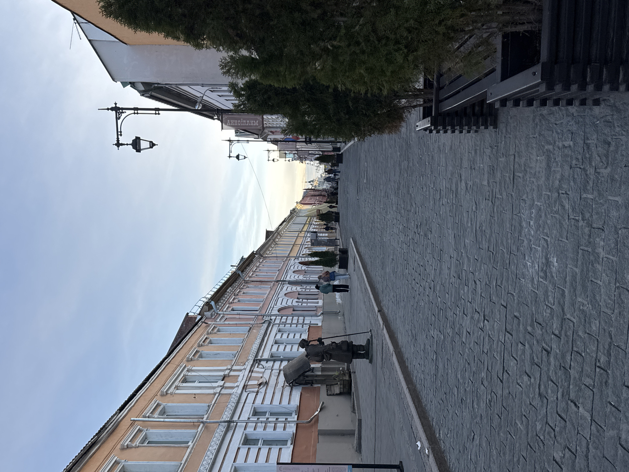 The image shows a street scene in Nizhny Novgorod, Russia. The road is paved with cobblestones. Buildings line the left side of the street; they are multi-story and feature decorative window frames, a light tan hue on the exterior walls, and white trim. People are walking along the sidewalk, including a statue. The sky is partially overcast with the setting sun in the distance. Streetlights line the road.