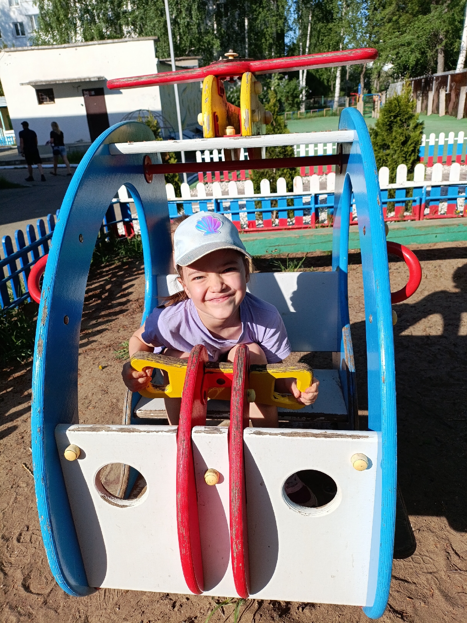 The image captures a young girl with brown hair, wearing a light purple shirt and a light blue cap adorned with a seashell graphic, seated in a playground structure resembling a helicopter. She is smiling broadly at the camera, holding onto a yellow and red handlebar.
The structure is painted in bright primary colors: blue arches frame the sides, with a white front panel featuring circular openings. A red and yellow propeller-like fixture sits atop the structure. The ground around the helicopter is sandy.
In the background, a building with a light-colored facade is visible, along with a section of a colorful fence painted in blue, white, and red. Two people, possibly adults, are walking away in the distance. Green trees are present as well.
The photograph was taken in Chaikovsky, Russia.