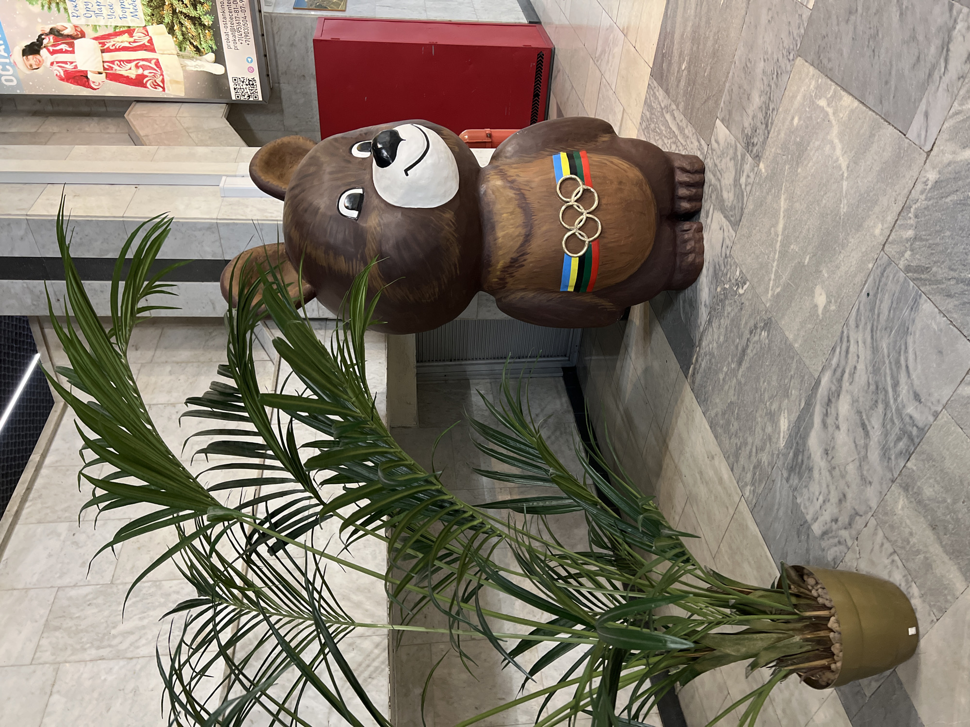 The image captures an indoor scene, likely in a public building such as a mall or lobby, in Khimki, Russia. The central subject is a large, brown statue of Misha, the bear mascot of the 1980 Moscow Olympics. Misha features a white face, black nose, and eyes, and wears a belt with the five Olympic ring colors (blue, yellow, black, green, red) underneath the Olympic rings themselves.
To the left of the bear, a tall, vibrant green palm-like plant sits in a dark pot. The floor is covered with a patterned arrangement of light and dark grey marbled tiles. In the background, a large poster on a light wall features a smiling woman dressed as a Snegurochka (Snow Maiden). Visible Cyrillic text on the poster, partly deciphered by OCR, includes "ОСТАНКИНО" (Ostankino) and words that appear to be categories like "Пар" (possibly "Парик" for wig), "Усы" (Moustache), "Боро" (likely "Борода" for beard), and "Мебе" (likely "Мебель" for furniture), suggesting a rental service. Contact information such as "prokat-ostankino", an email, and Russian phone numbers "+7(495)617-81-0" and "+7(903)504-07-9" are also visible, along with a QR code. Behind the bear, a red rectangular object, possibly a utility box or cabinet, is positioned against the wall. The scene is brightly lit by indoor lighting, and the bear statue conveys a friendly, welcoming emotion.