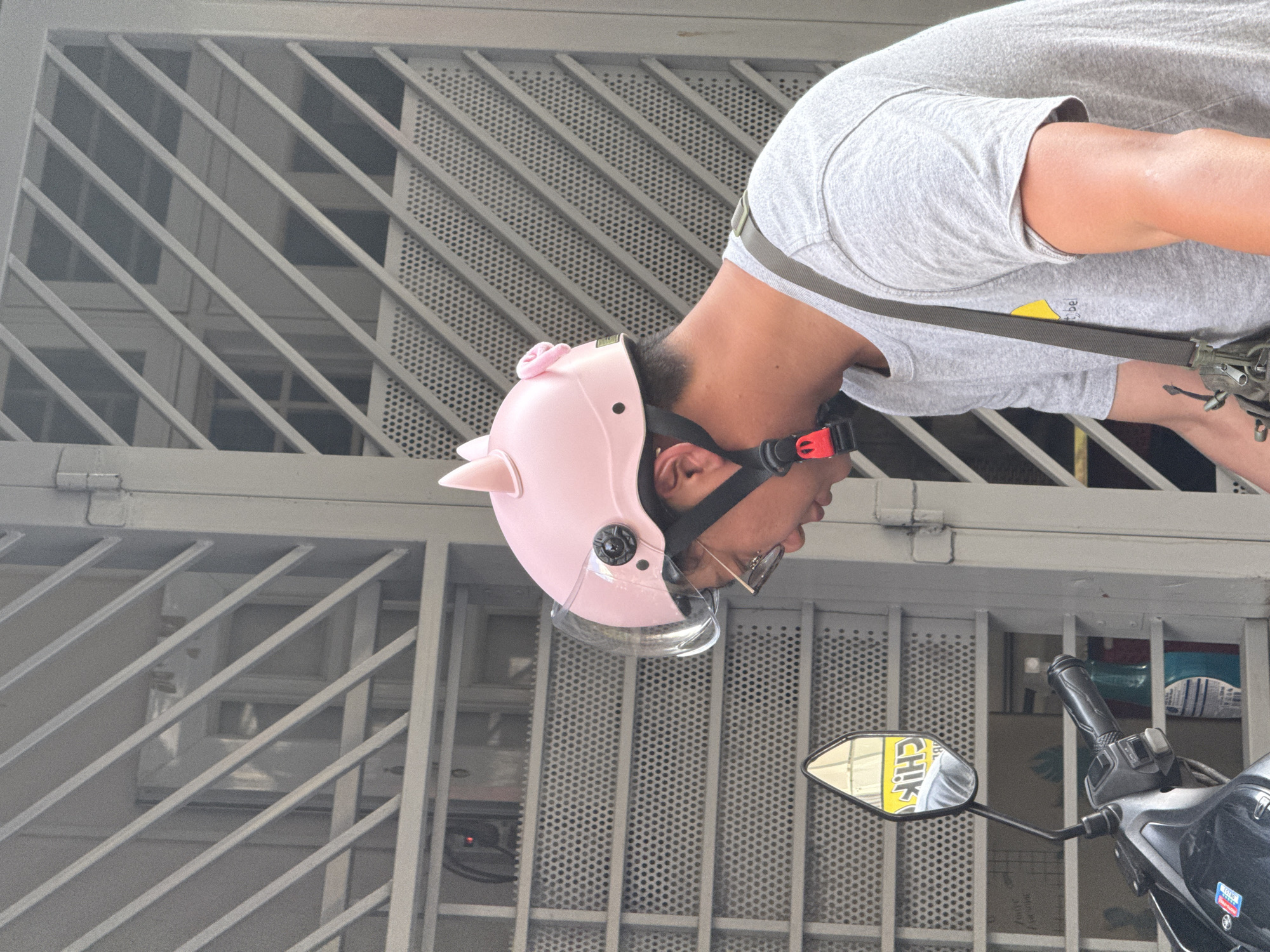 A person wearing a pink helmet with pig ears and a gray t-shirt is visible on a motorcycle. The motorcycle's side mirror has a reflection of a yellow sign with the text "CHIK." The backdrop consists of a gray metal grating, likely a security barrier on a building. The location appears to be in a densely populated area in Nha Trang Ward, Vietnam, evidenced by the architectural style and density of the buildings behind the grating. The orientation of the image suggests it was taken from a vertical perspective.