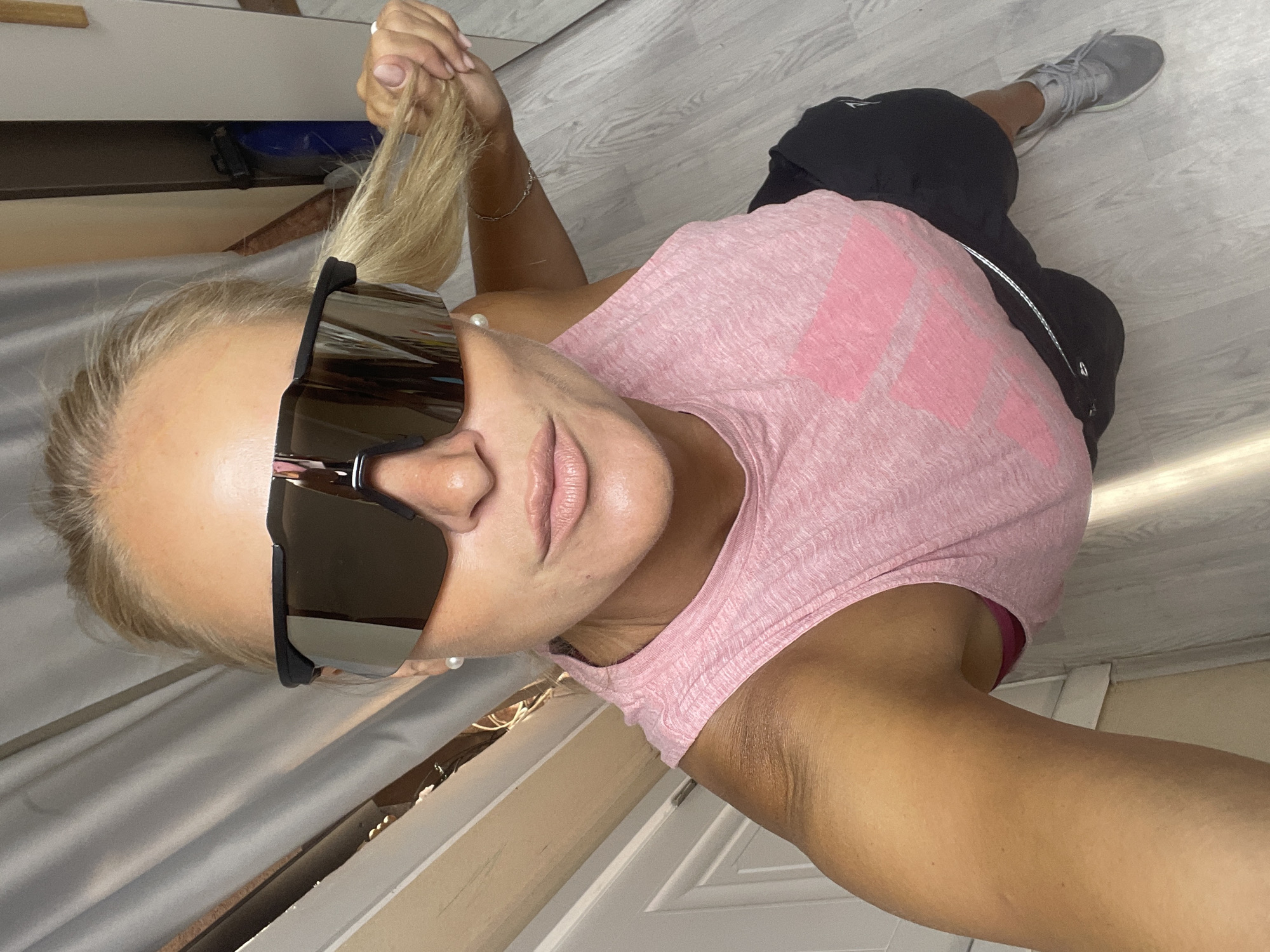 A woman with blonde hair pulled back is taking a selfie in what appears to be a home environment. She is wearing oversized, reflective sunglasses, a pink sleeveless tank top with a large pink logo, and black shorts. Her right hand is pulling her ponytail. The background shows a light-colored wooden floor, a white door, and a gray curtain. There are no indications that this scene is located in the city of Baranovka, Russia.