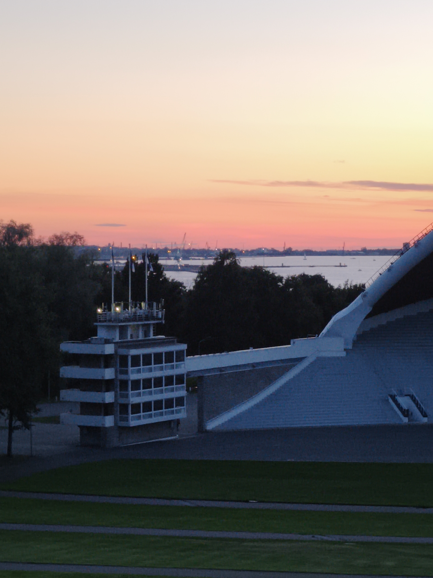 The image depicts a view of the Pirita beach and the adjacent area in Tallinn, Estonia, at sunset. In the foreground, a manicured green lawn with pathways is visible. Behind the lawn, a multi-story building with a white facade and numerous windows stands, topped with a structure featuring flags and lights, possibly a control tower or viewing platform. Further in the background, a vast body of water, likely the Baltic Sea, stretches towards the horizon. Along the coastline, trees and industrial structures, including cranes, are discernible, silhouetted against the colorful sunset sky, which transitions from pale yellow to pink and orange. The sky is partly cloudy, with thin clouds stretched across the horizon. The overall scene conveys a serene atmosphere characteristic of a seaside location during twilight.