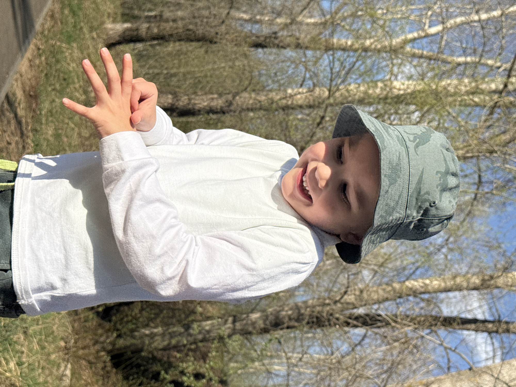 A young child is outdoors, wearing a white long-sleeved shirt and a gray camouflage bucket hat. The child is smiling and holding up four fingers. The location appears to be in a park with bare trees, possibly in early spring. The scene is sunny and bright, suggesting a clear day. The location in the city of Chaikovsky, Russia is not ascertainable.
