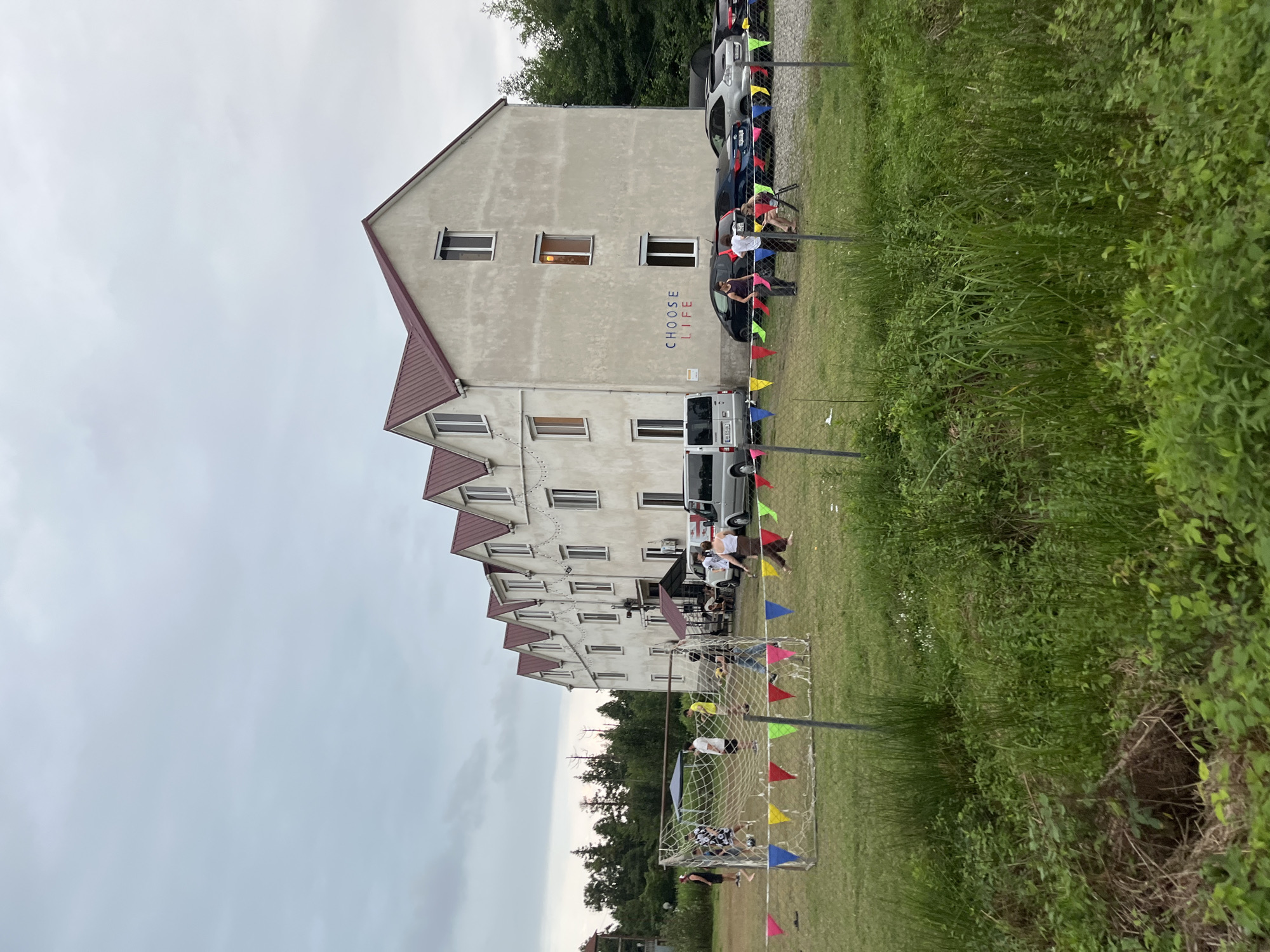 In Ureki, Georgia, a building with a red-tiled roof and a light-colored facade features the words "CHOOSE LIFE" painted in blue and red on its exterior. The building has multiple windows and appears to be a multi-story structure. In front of the building, a grassy field has a soccer goal set up, and several people are engaged in a soccer game. Colorful triangular flags are strung between posts in front of the soccer field. Several cars are parked in front of the building. The environment is outdoors with trees visible in the background and tall green grass in the foreground. The sky is overcast.