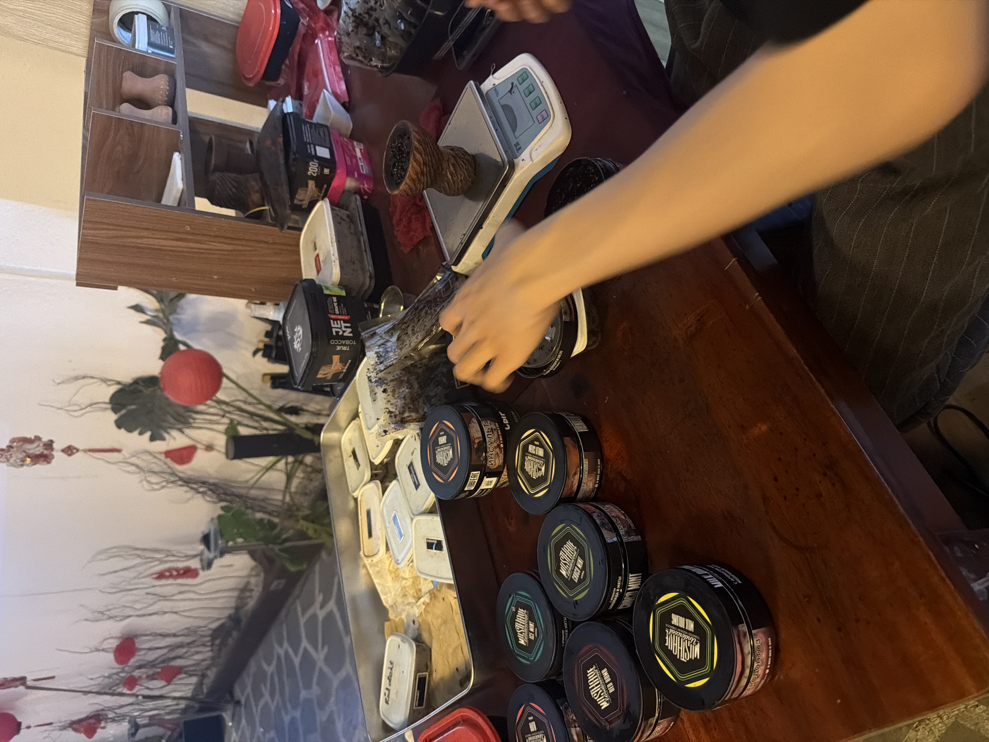 The image shows a person preparing hookah tobacco. A person's hand is reaching towards a tray filled with multiple round containers of MUSTHAVE brand hookah tobacco, including flavors such as "Red Bomb," "Ice Mint," and "Choco Mint." There is also a package of "True Tobacco Grape Me" visible. The tobacco is being weighed on a digital scale next to a hookah bowl. The background includes decorative elements like a red paper lantern and some artificial foliage. The presence of hookah tobacco suggests this might be in a cafe or lounge in a place in Vietnam, perhaps in Đà Nẵng.