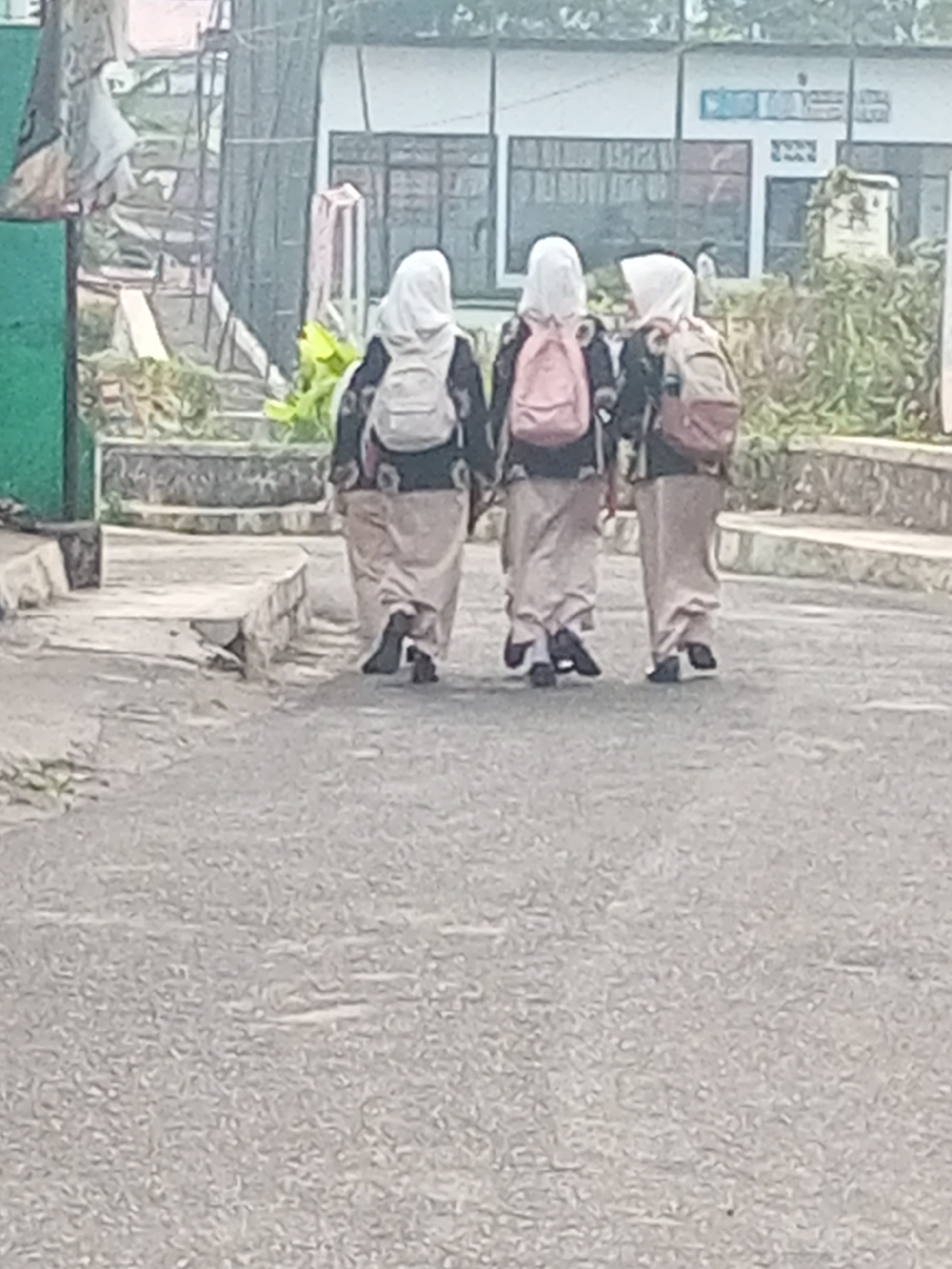 In Krasak, Indonesia, three individuals wearing hijabs and school uniforms walk away from the camera on a paved surface. They each carry a backpack. The person in the middle has a pink backpack, while the person on the right has a brown backpack. The person on the left's backpack is partially obscured. In the background, there is a building with windows and some greenery. The overall lighting suggests it is daytime.