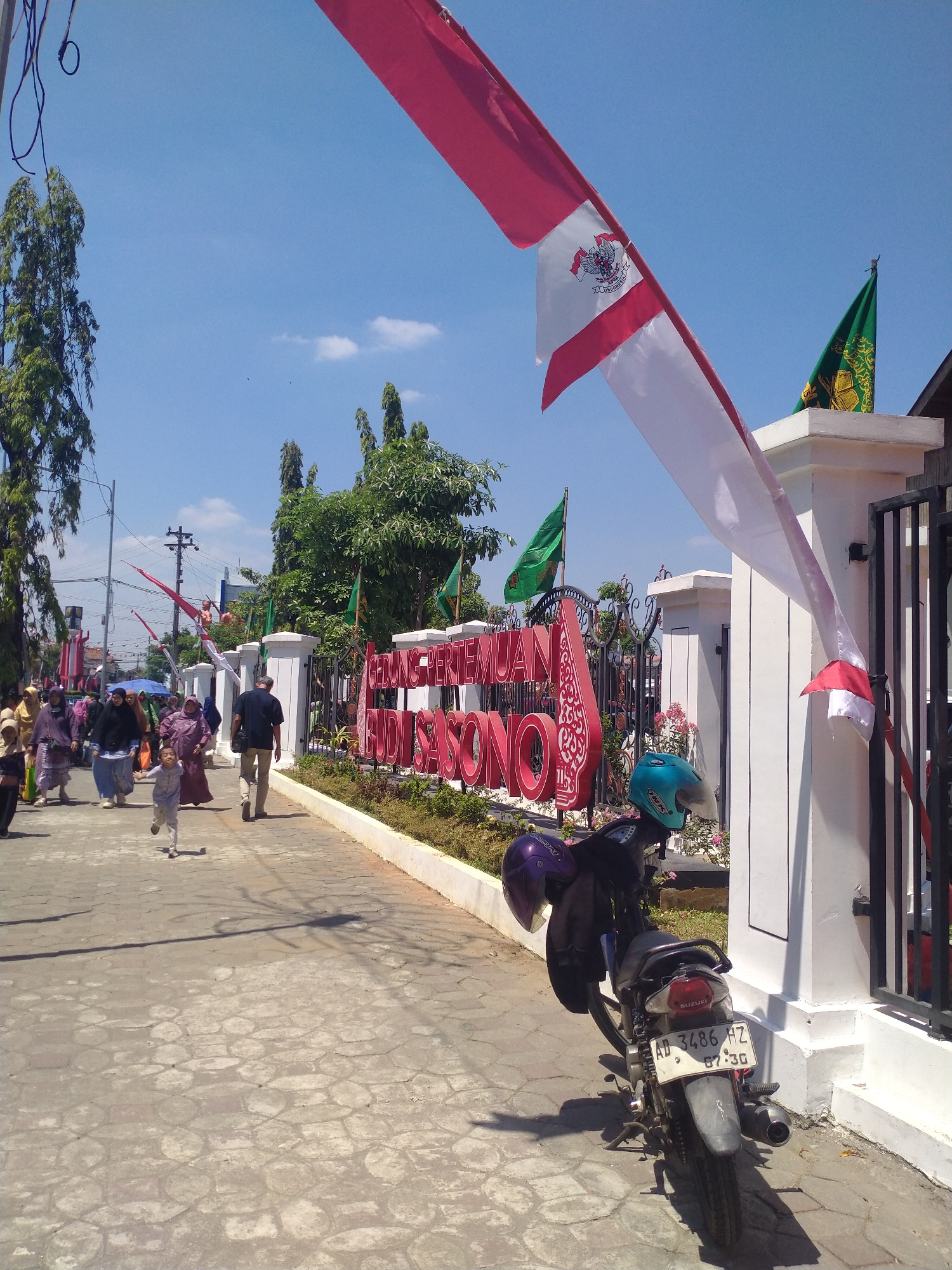 The image depicts a street scene in front of a building with large red lettering that reads "GEDUNG PERTEMUAN BUDI SASONO," located in Sukoharjo, Indonesia. The building is adorned with Indonesian flags, both red and white, and green flags with yellow insignia. A paved walkway lines the front of the building, bordered by a low wall and greenery.
Several people are present on the walkway. In the foreground, a child in a light-colored outfit runs towards the viewer. Further back, a group of individuals, mostly women wearing headscarves and modest clothing, are walking along the path. A man in a dark shirt and beige pants walks away from the viewer.
A motorcycle is parked on the right side of the walkway, facing away from the viewer. It has two helmets secured to it, one blue and one purple. The license plate of the motorcycle is visible, displaying "AD 3486 HZ" and "07.30".
The sky is clear and blue with a few scattered white clouds. Trees with lush green foliage are visible on the left side of the image and behind the building. The overall impression is a bright, sunny day in an outdoor public space.