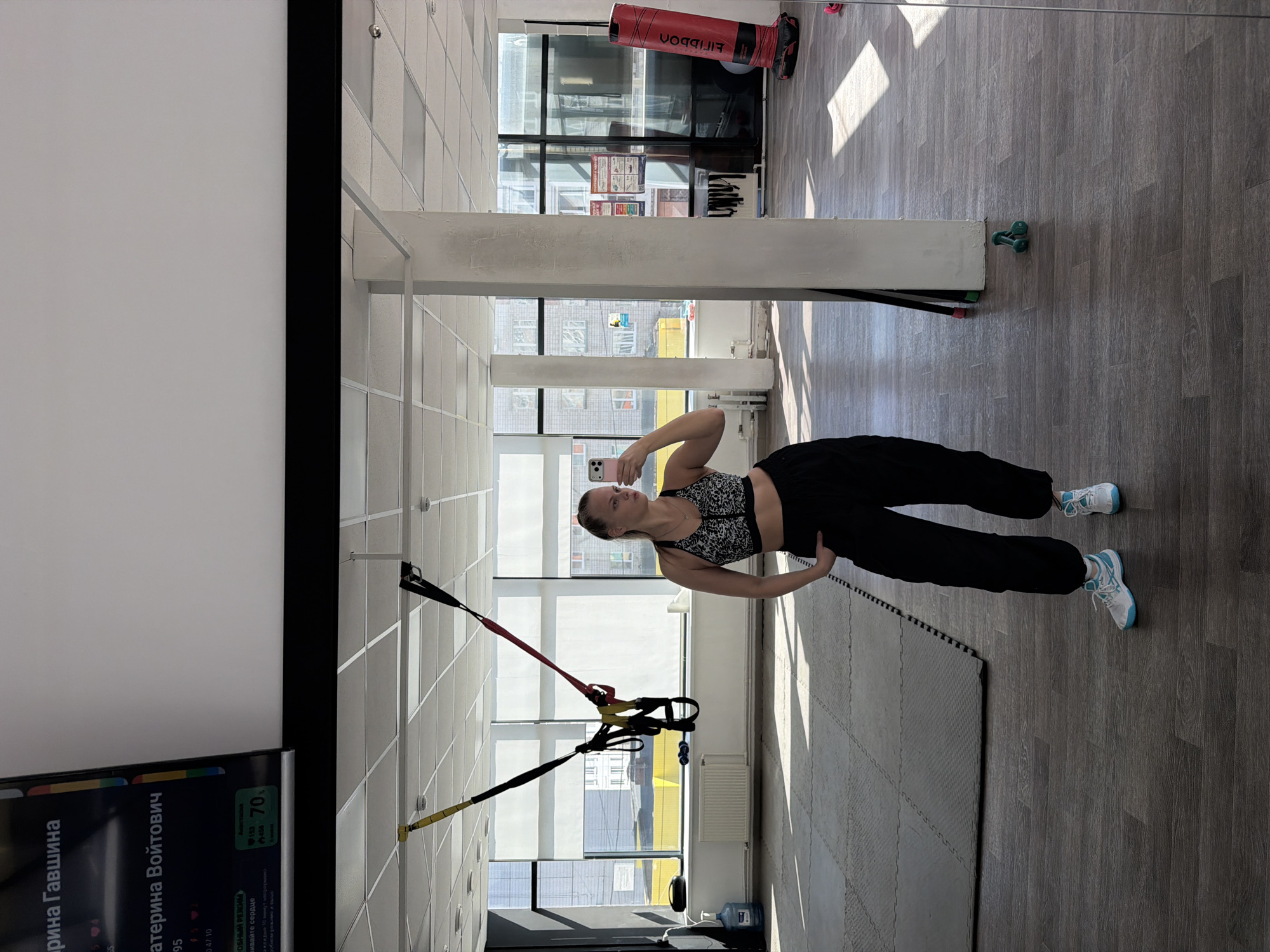 An indoor scene, likely a fitness studio or gym in Izhevsk, Russia, is depicted during what appears to be daytime, illuminated by bright natural light. The primary subject is a woman, captured in a mirror selfie, showing her reflection from the torso up. She has light hair, pulled back, and is wearing a black and white leopard-print sports bra paired with black athletic pants. She holds a pink-cased smartphone in her hand to take the photograph.
In the background, large windows reveal an urban environment with buildings outside. A TRX suspension trainer system, featuring yellow, red, and black straps, hangs from the ceiling. A red punching bag, labeled "FIILDDON", is partially visible further back. The floor is made of wood-patterned laminate, with a black exercise mat laid out in the foreground. A green dumbbell or similar fitness accessory is situated on the floor near a white column, and a large water dispenser bottle is also present.
To the left of the image, a large screen displays various details in Russian. Visible text includes "рина Гавшина", "катерина Войтович", along with numbers such as "95", "47 10", "153", and "70". Phrases like "-ОБНЫЙ РЕЖИМ" (likely part of "тренировочный режим" - training mode) and "живайте сердце" (likely part of "отслеживайте сердце" - track heart) are also discernible, suggesting fitness tracking information. The overall setting is well-lit and equipped for various physical activities.