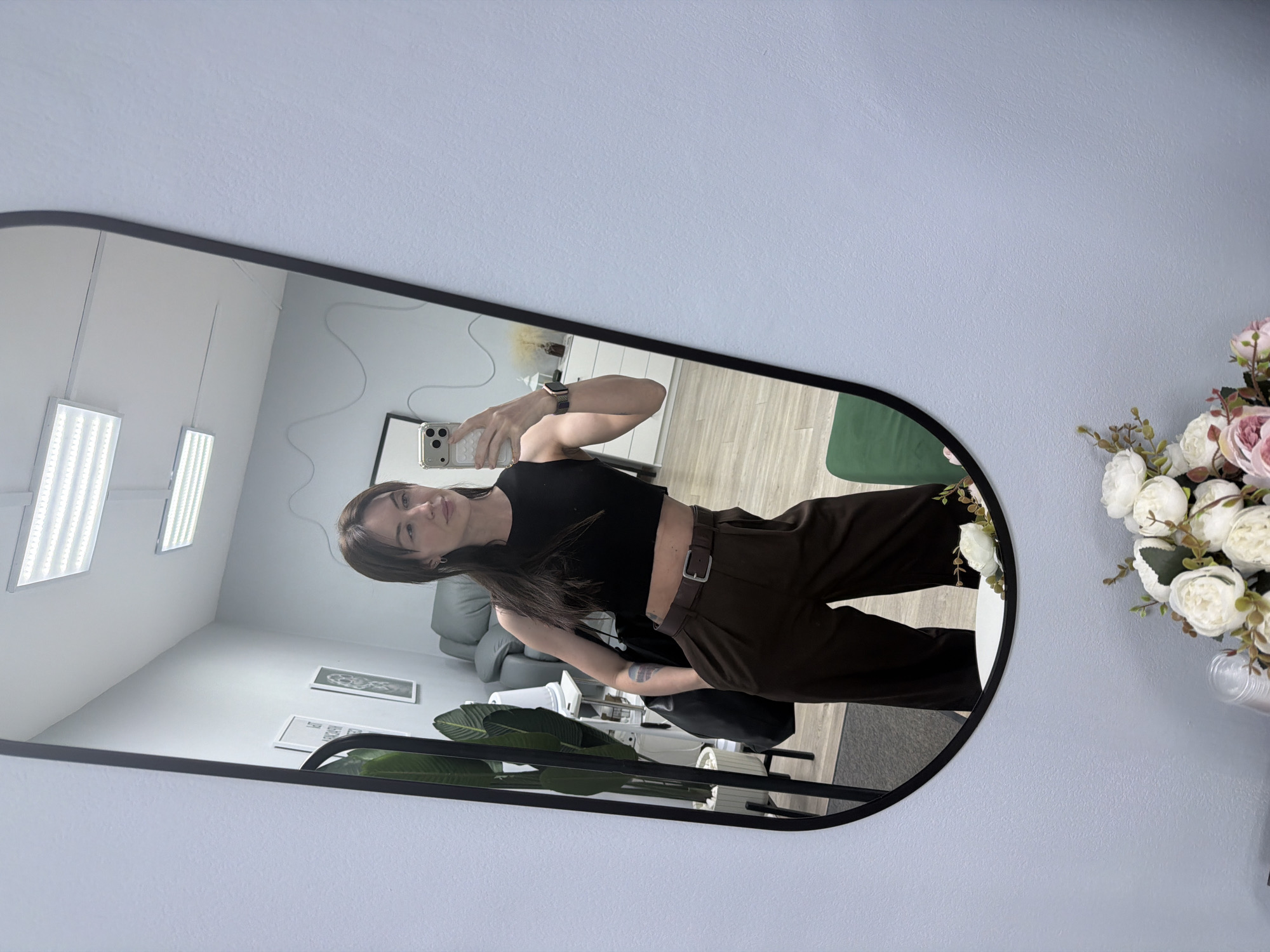 A young woman stands in front of a mirror, taking a selfie in what appears to be a room in Perm, Russia. The woman is wearing a black crop top and brown pants with a belt. In the mirror's reflection, there are white rectangular lights, two framed pictures on the wall, a plant in a pot, and a gray couch. To the right of the mirror are pink and white flowers.