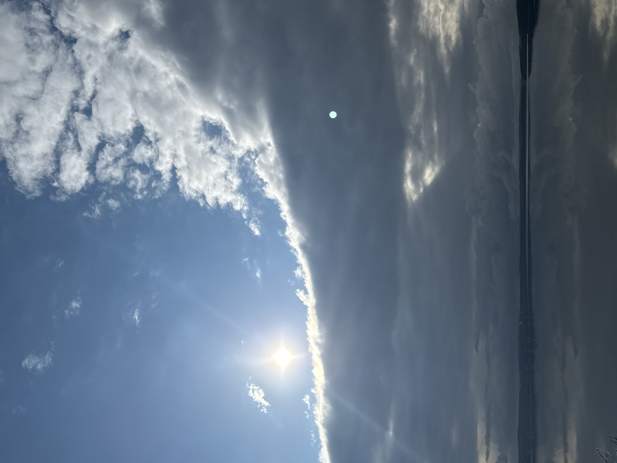 The image captures a dramatic sky scene, dominated by contrasting weather conditions. The left half of the frame features a bright, clear blue sky with scattered white, wispy clouds. A brilliant sun shines intensely in this clear expanse, emanating prominent rays of light.
To the right, a significant portion of the sky is covered by dense, dark grey clouds, creating a stark visual divide from the sunny area. The border where the bright blue sky meets these dark clouds is illuminated, highlighting the edges of the cloud formations. Within the darker cloud mass, a small, distinct light blue or white circular spot is visible, likely a lens flare.
Along the far right edge of the image, a long, narrow body of water is present, acting as a reflective surface for the dark, cloudy sky above. The water shows elongated, vertical reflections of the clouds, with subtle variations in light and shadow. A faint, dark strip running vertically along the water's edge suggests a distant shoreline or landmass.
The scene depicts a dynamic interplay of light and shadow under a partially cloudy sky during daylight hours, likely mid-day due to the sun's intensity. There are no people or discernible text. The natural landscape, featuring a vast sky and a body of water, evokes a sense of environmental grandeur, possibly near the Votkinsk Reservoir in Russia.