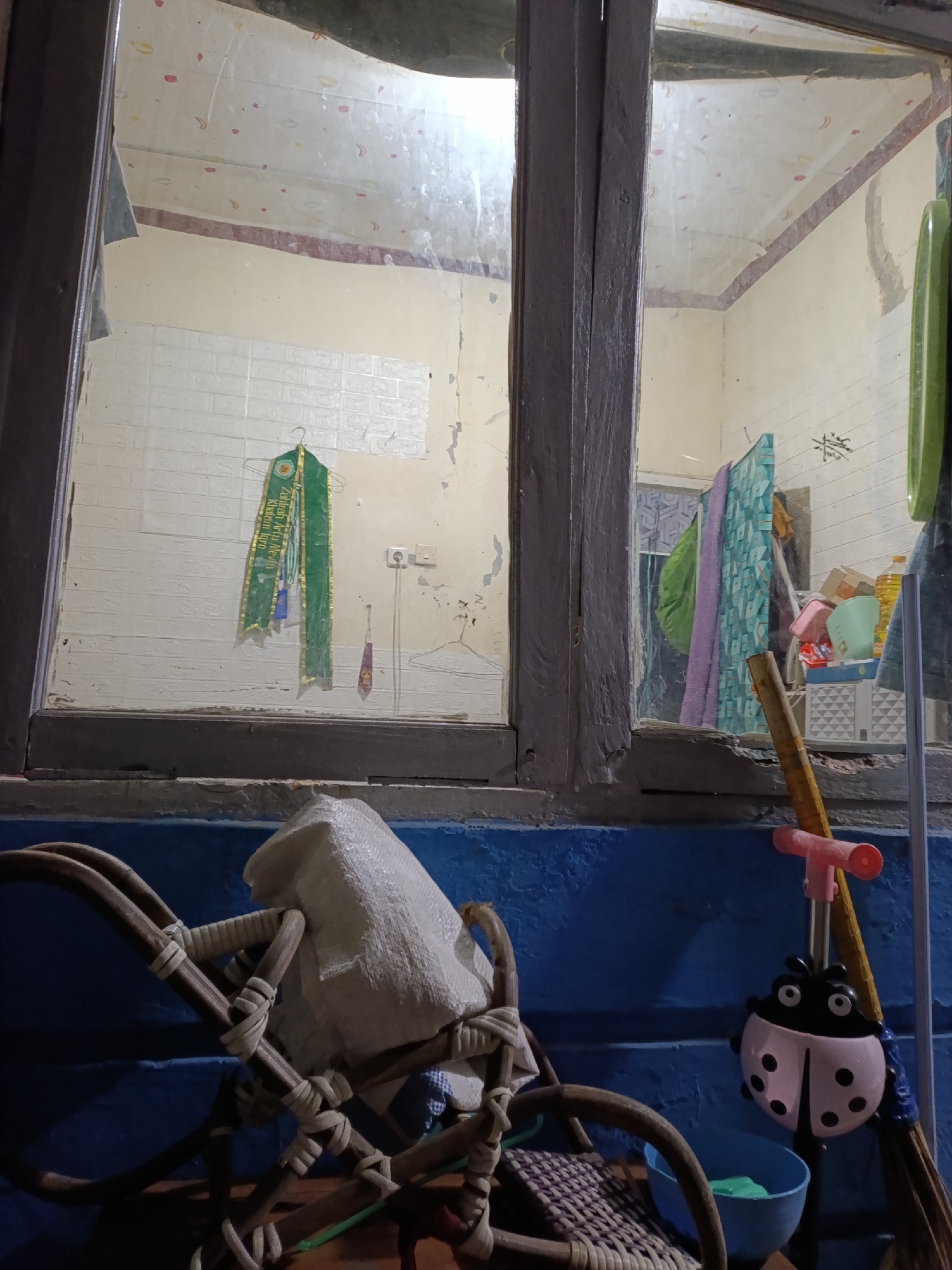 The image depicts an interior view through a dirty window in Cariu, Indonesia. The room visible through the window is sparsely furnished and appears somewhat disheveled. The walls are cream-colored, with some areas showing peeling paint. There is a green sash with gold trim hanging on a wall covered in white, textured tiles. A small purple object hangs from a hook to the right of the sash. The ceiling is visible at the top, with a patterned wallpaper. Various items are placed on shelves or the floor in the background, including what appears to be fabric, a plastic container, and a bottle of liquid.
In the foreground, outside the window, part of a woven chair is visible, covered with a white sack. To the right, a broom with a pink ladybug-shaped accessory is leaning against the wall. A blue bowl is situated near the base of the broom. The overall impression is of a humble, possibly lived-in space.
